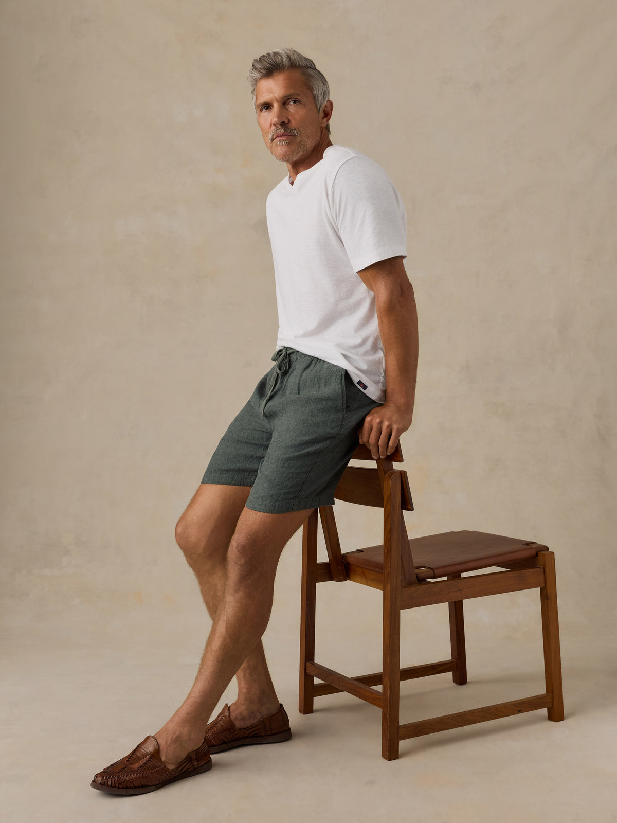 A man wearing a white short-sleeve polo shirt and dark green drawstring shorts leans against a wooden chair in a studio setting with a textured beige background., image 5 of 5