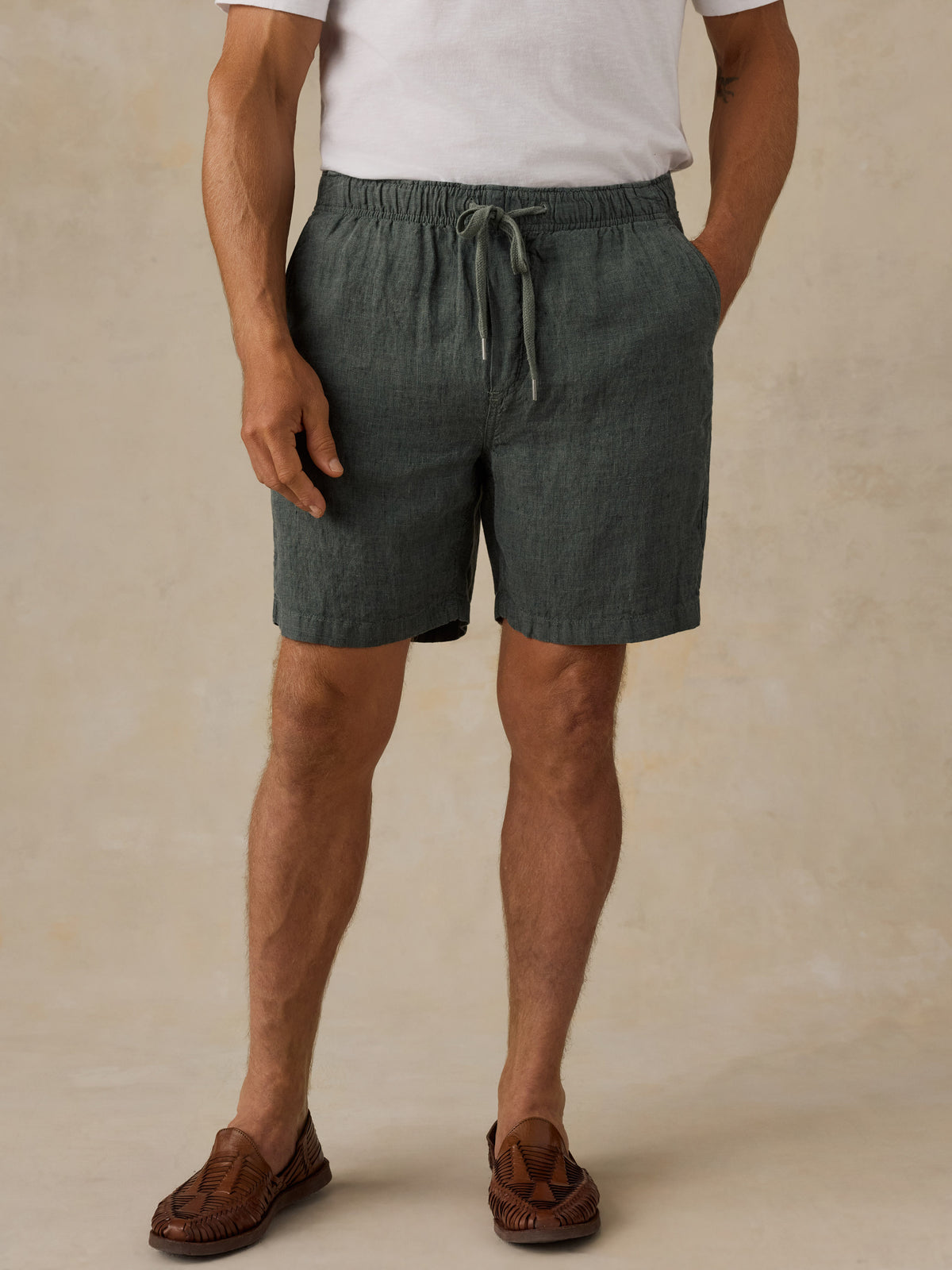 A man wearing dark green linen drawstring shorts, a white t-shirt, and brown leather loafers, standing against a textured beige studio backdrop., image 1 of 5