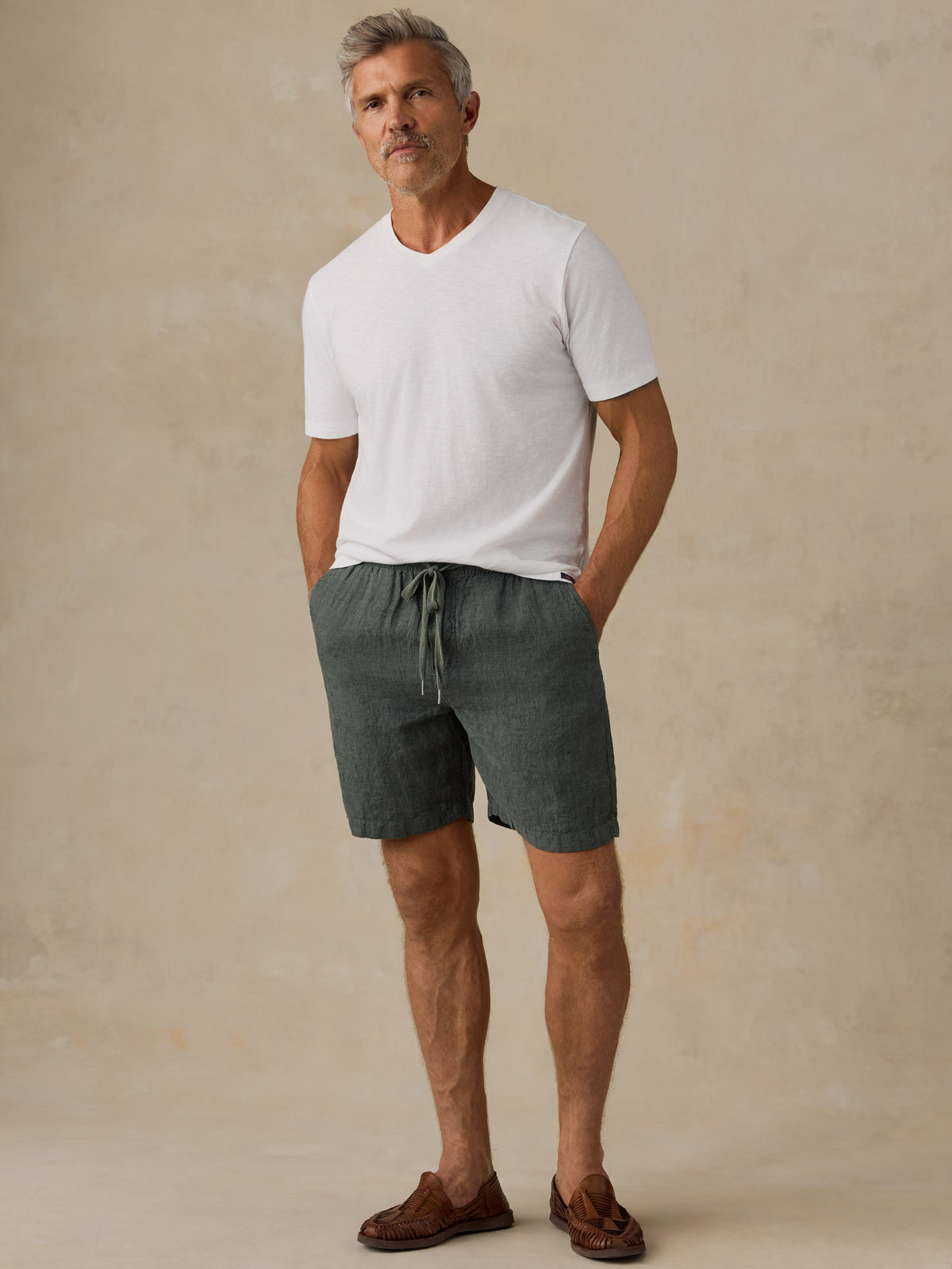 A man wearing a white short-sleeve V-neck t-shirt, dark green drawstring linen shorts, and brown leather loafers, standing against a textured beige studio backdrop., image 2 of 5
