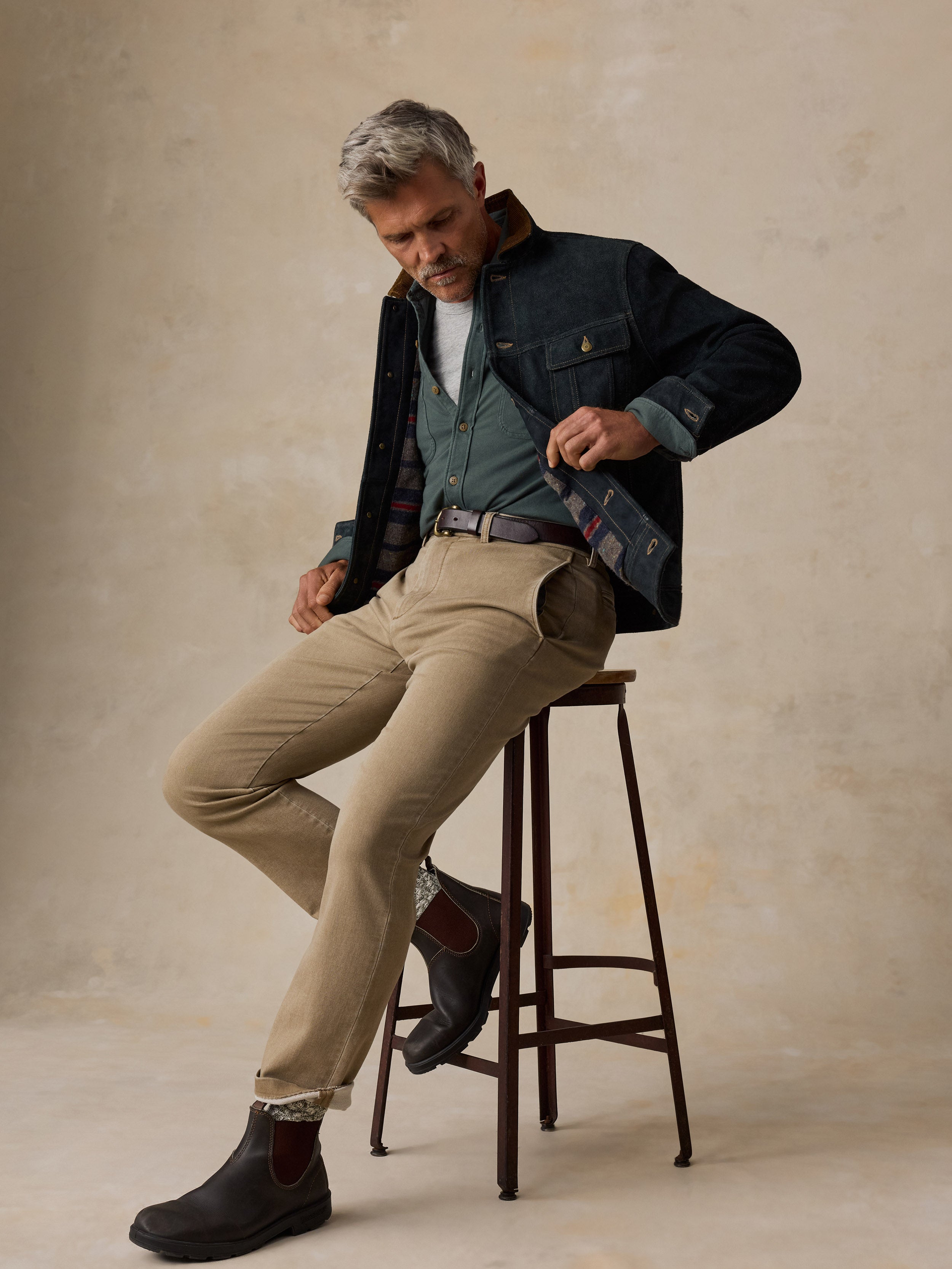 A man with gray hair sits on a dark wooden stool, wearing a layered outfit consisting of a dark navy overshirt, a green button-down shirt, a white t-shirt, beige trousers, and dark brown Chelsea boots, against a textured beige studio backdrop.