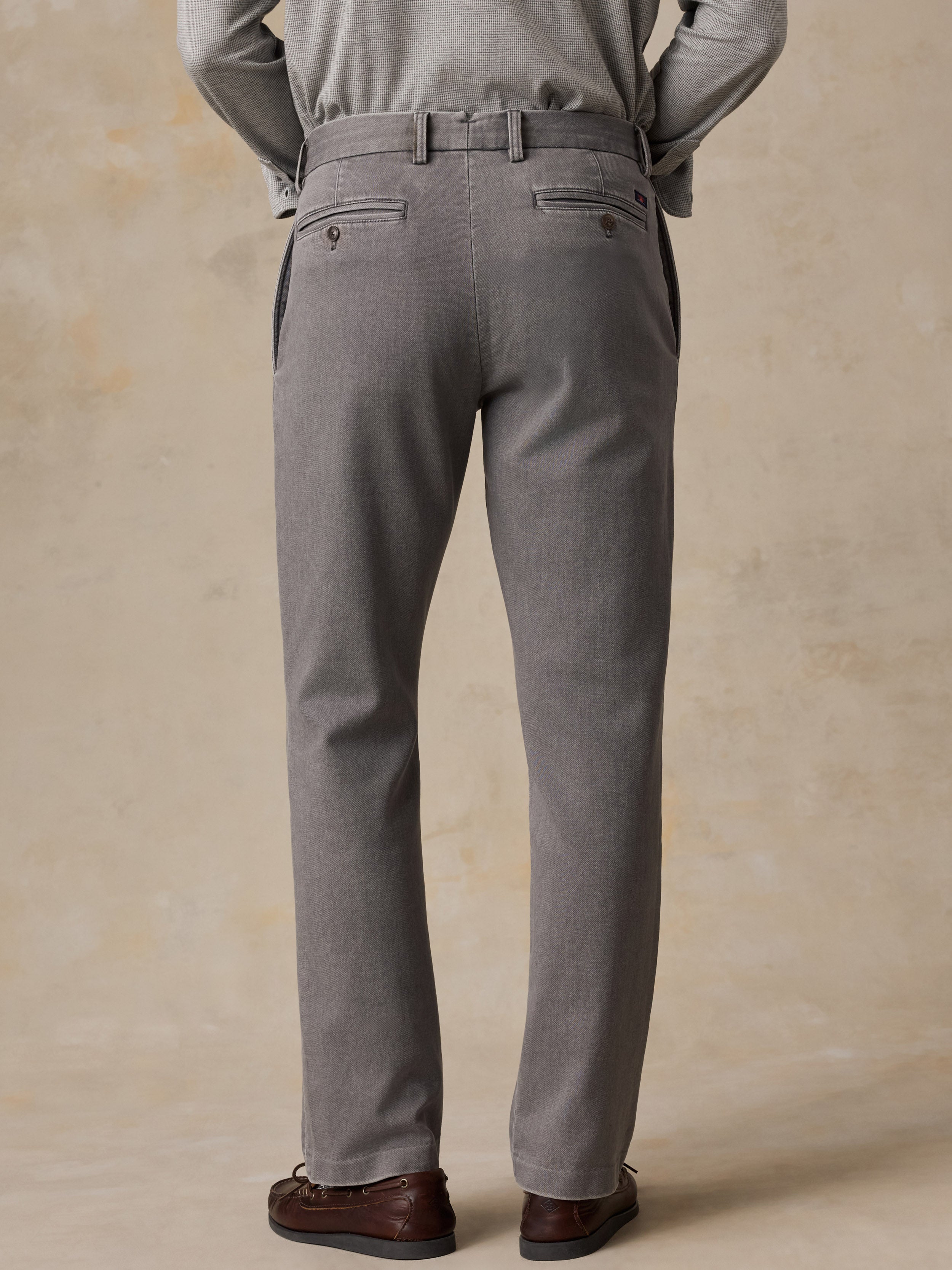 A man shown from the back wearing gray trousers with a matching belt, a light gray long-sleeve top, and dark brown loafers, standing against a textured beige studio backdrop.