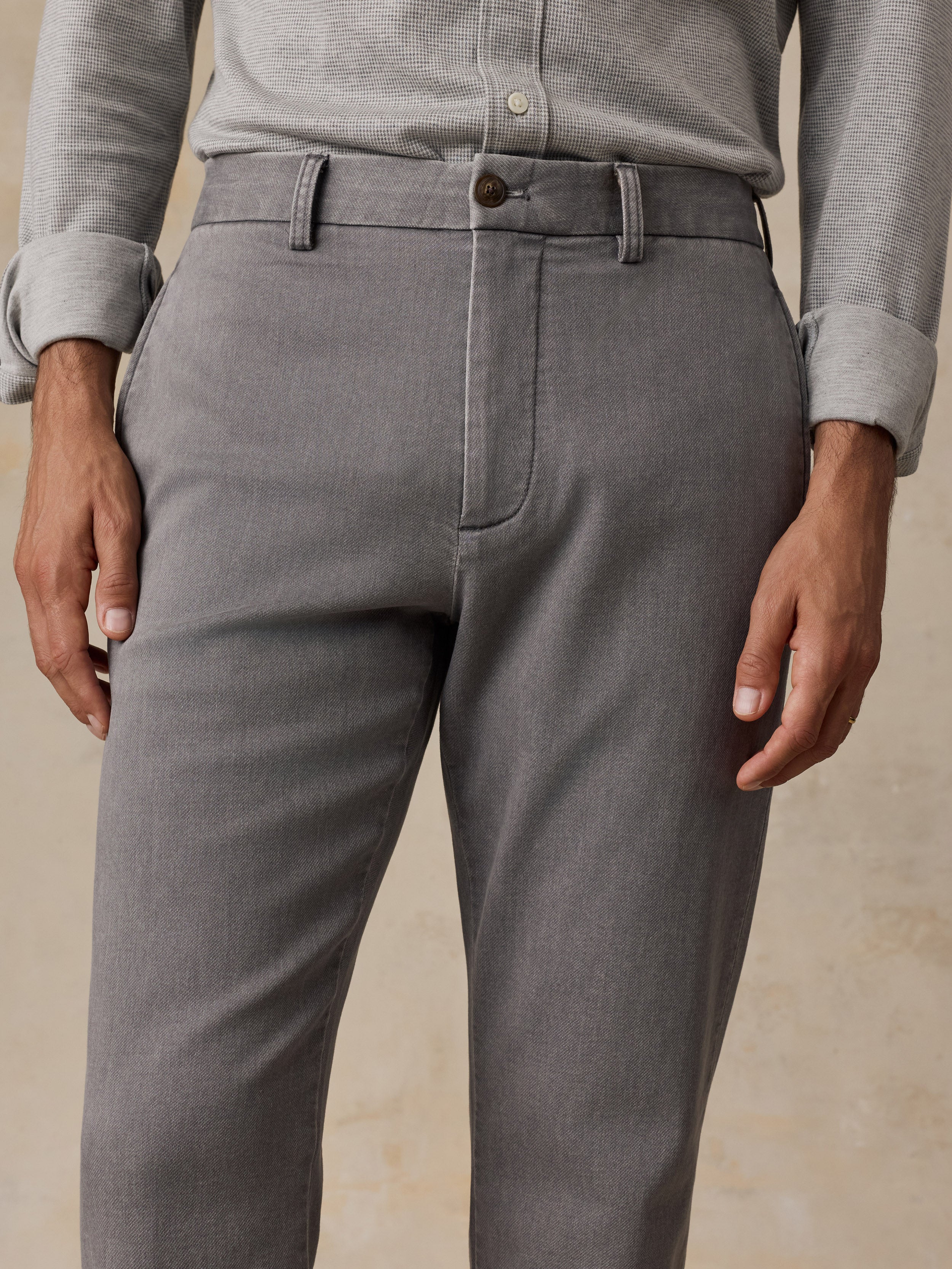 A close-up of a man wearing gray trousers with a matching gray belt, paired with a light gray button-up shirt, against a neutral beige background.