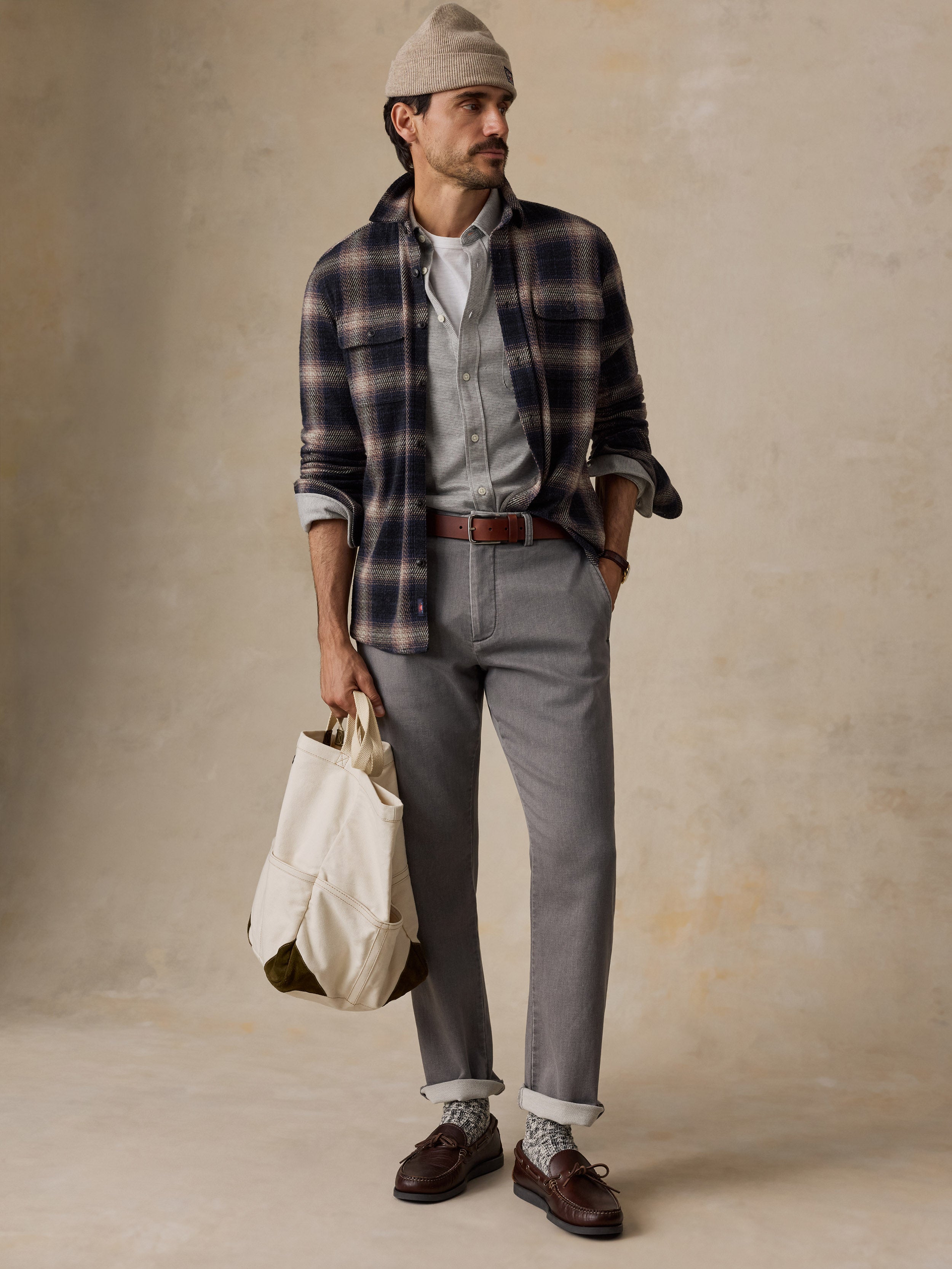 A man stands against a textured beige studio wall wearing a beige knit beanie, a navy and brown plaid flannel shirt over a light gray button-down, gray trousers, a brown belt, and dark brown loafers. He is holding a cream canvas tote bag.