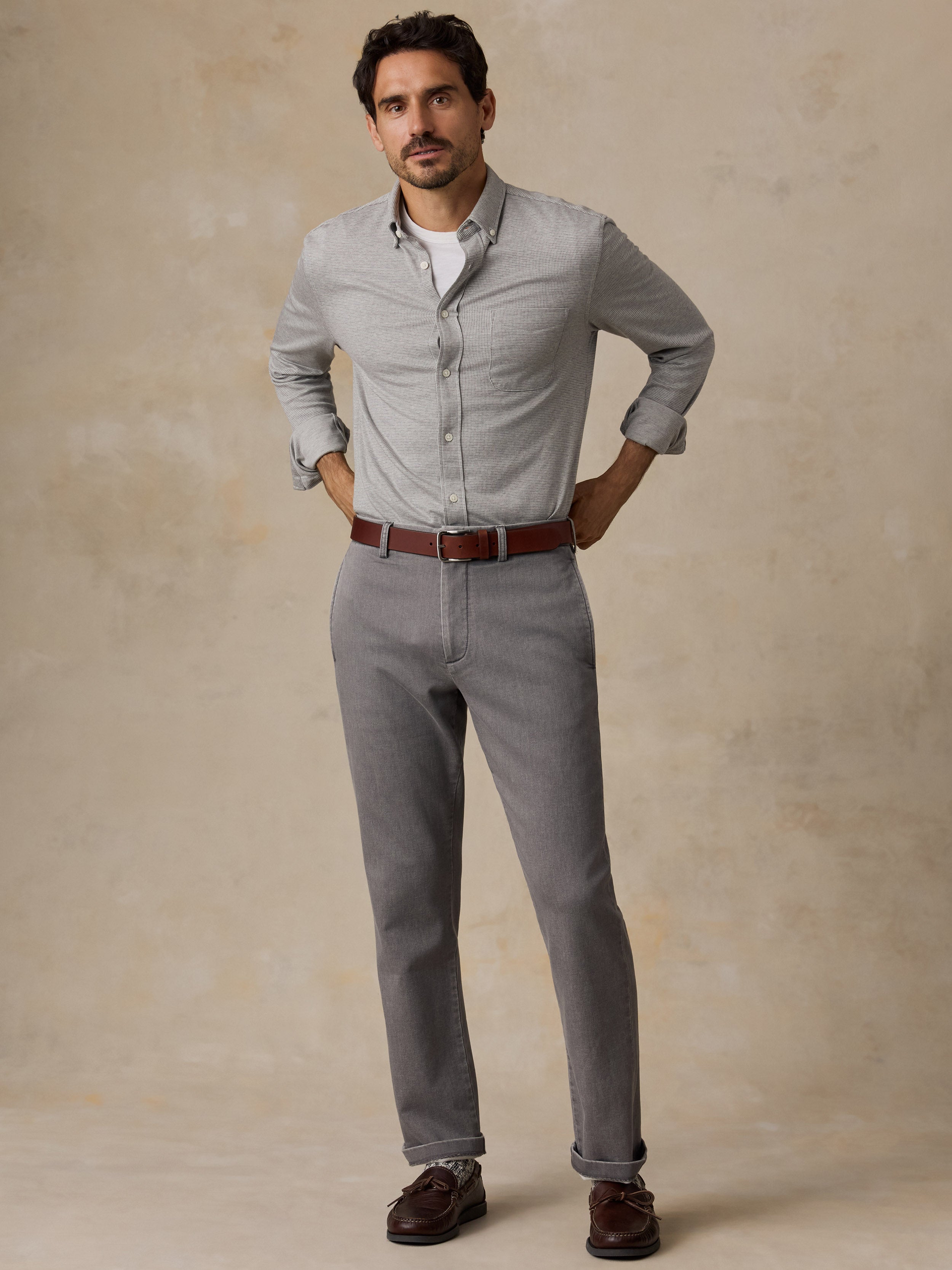 A man modeling a light gray patterned button-down shirt, gray trousers, a brown belt, and brown shoes against a textured beige studio backdrop.