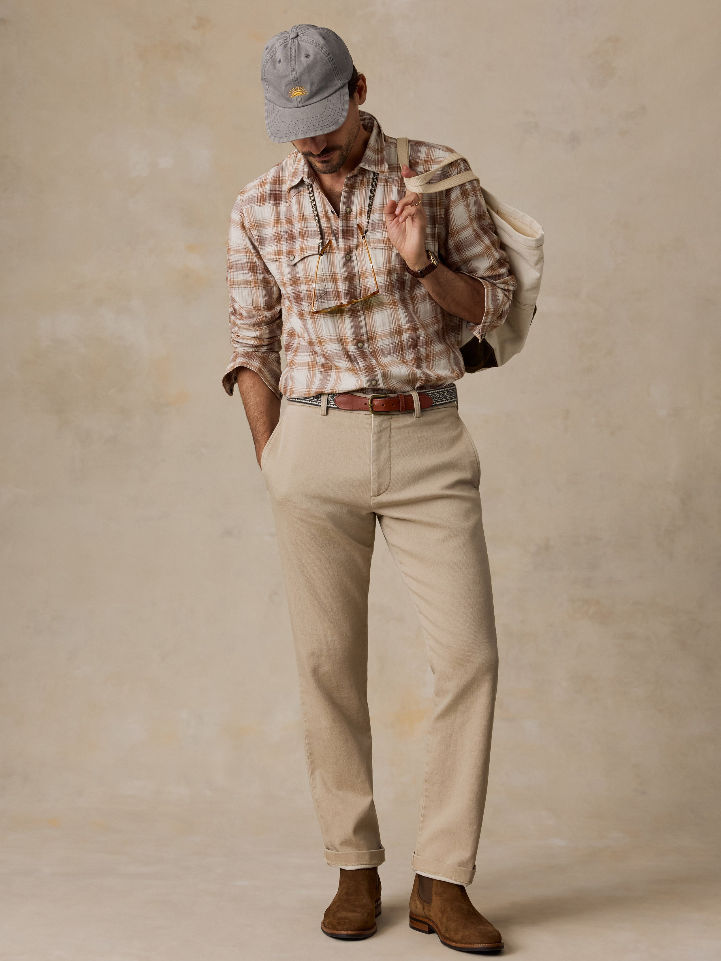 A man wearing a beige and brown plaid button-down shirt, khaki trousers, a brown belt, brown suede boots, a gray baseball cap, and carrying a canvas shoulder bag, standing against a textured beige studio backdrop.