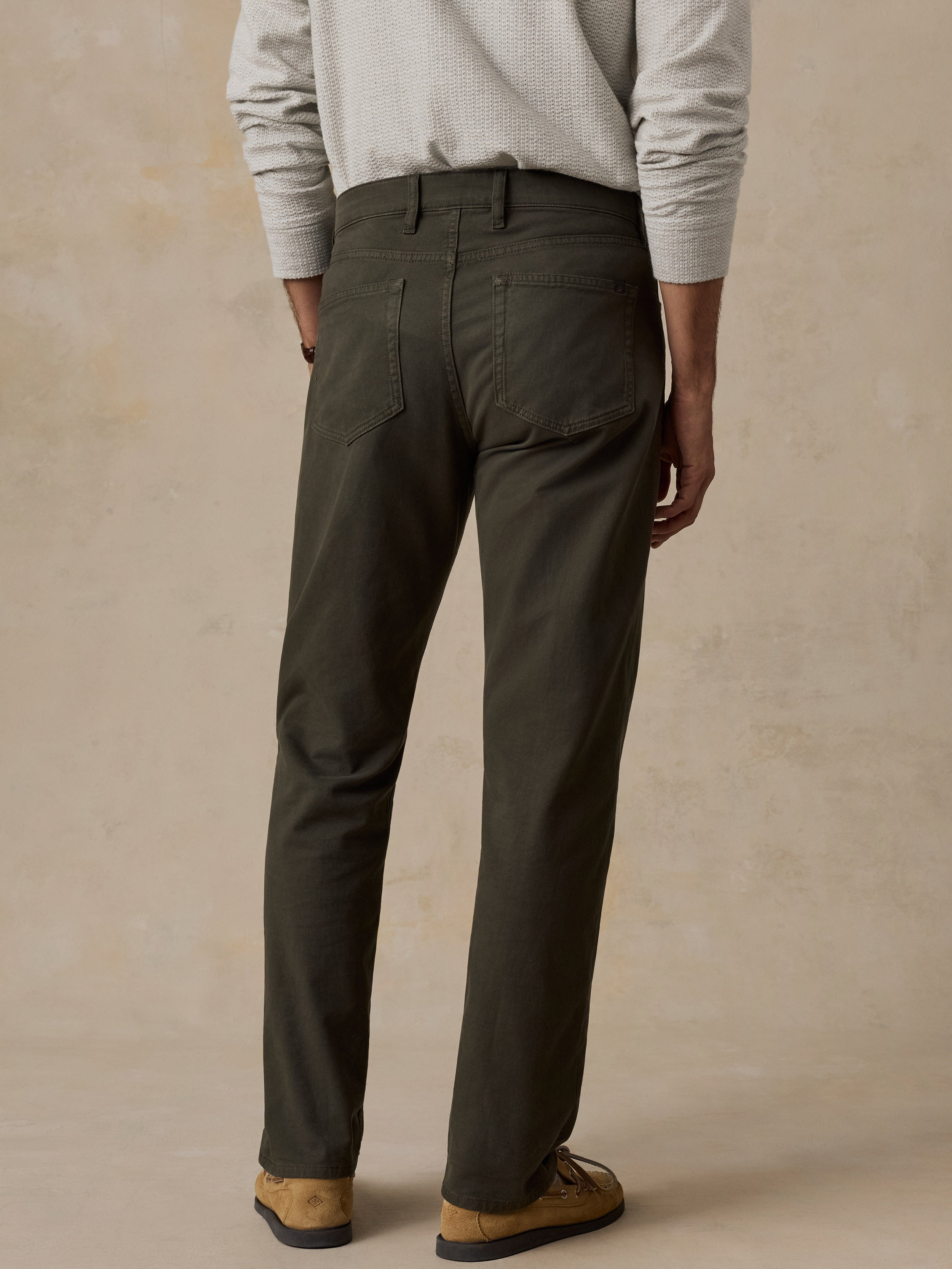 A person shown from the back wearing dark olive green straight-leg trousers, a light gray textured long-sleeve shirt, and tan suede loafers, set against a warm beige textured studio backdrop.