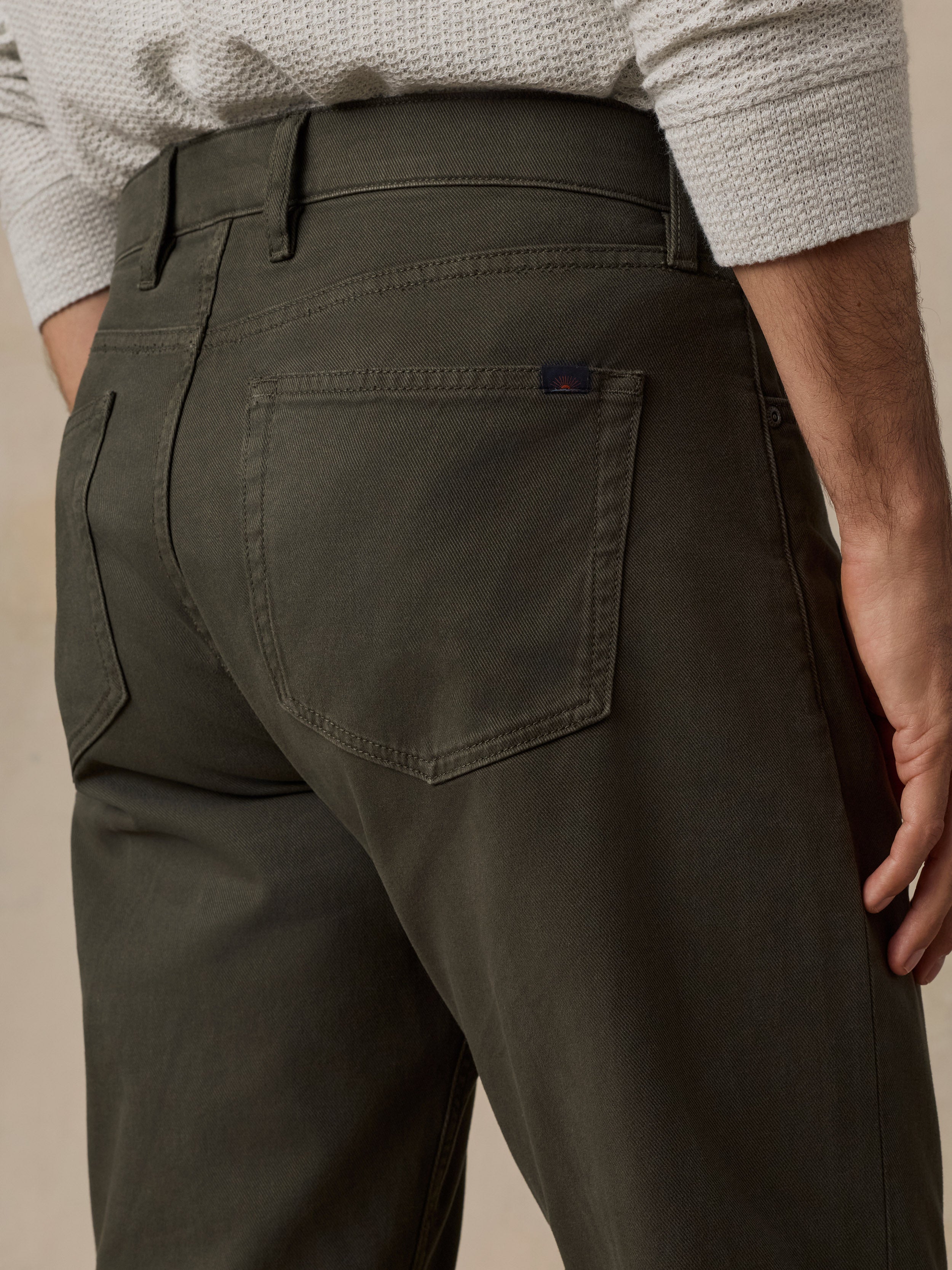 Close-up back view of a person wearing dark olive green trousers, showing the waistband, belt loops, back pockets, and a small blue brand tab on the rear right pocket.
