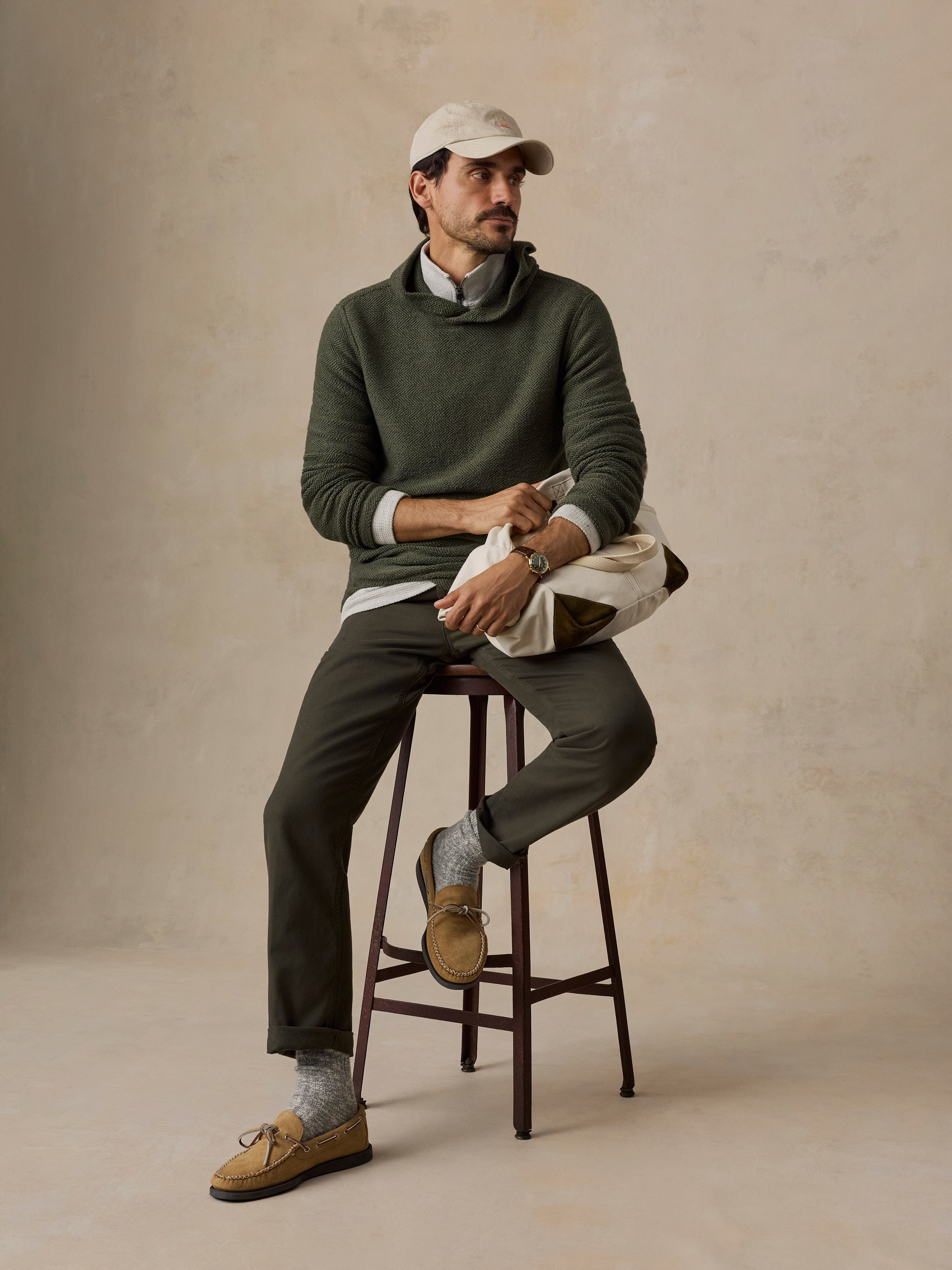 A man sits on a dark wooden stool wearing a dark green knit sweater over a white collared shirt, dark olive trousers, tan suede loafers, and a light beige baseball cap. He is holding a cream-colored knit bag against a textured beige studio backdrop.