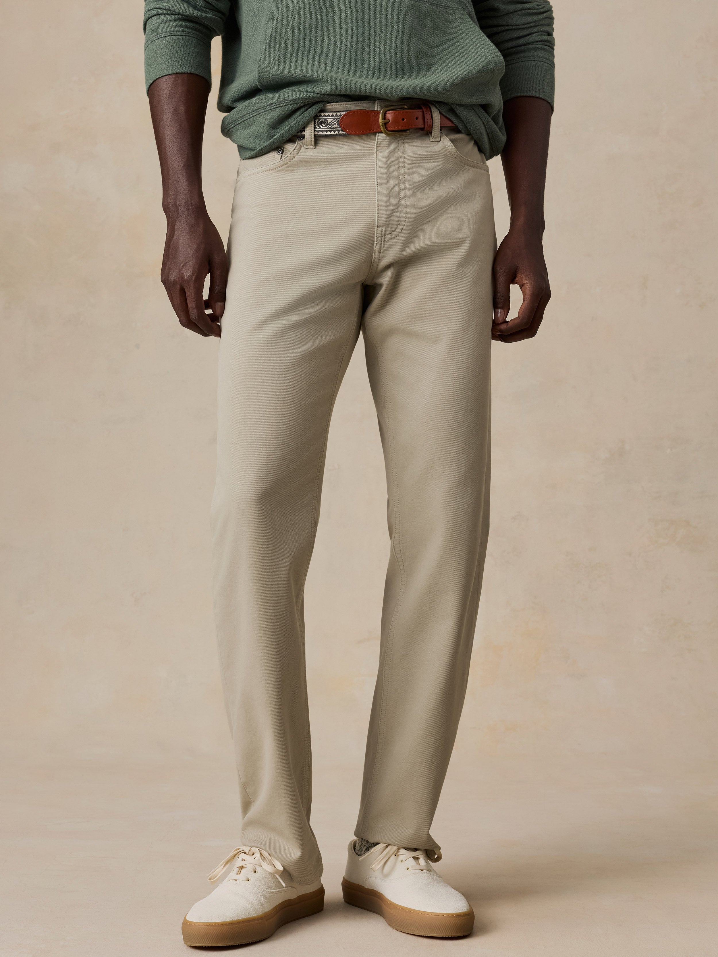 A model wearing a pair of light beige chinos with a brown leather belt, styled with a sage green long-sleeve shirt and off-white sneakers, set against a textured beige studio backdrop.