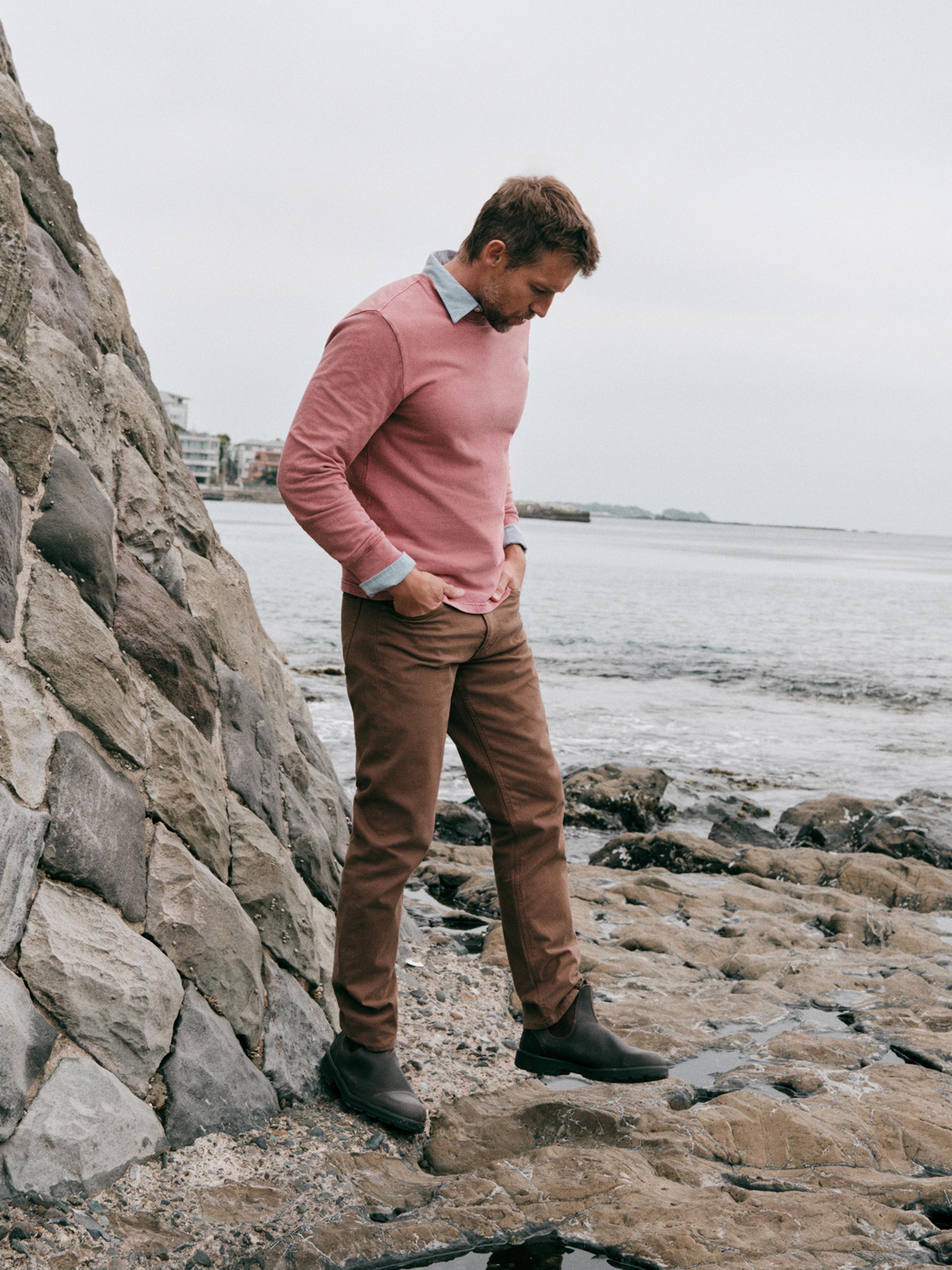 A man wearing a dusty pink long-sleeve sweater, brown trousers, and dark boots stands on a rocky shoreline next to a stone wall, looking down toward the rocks.