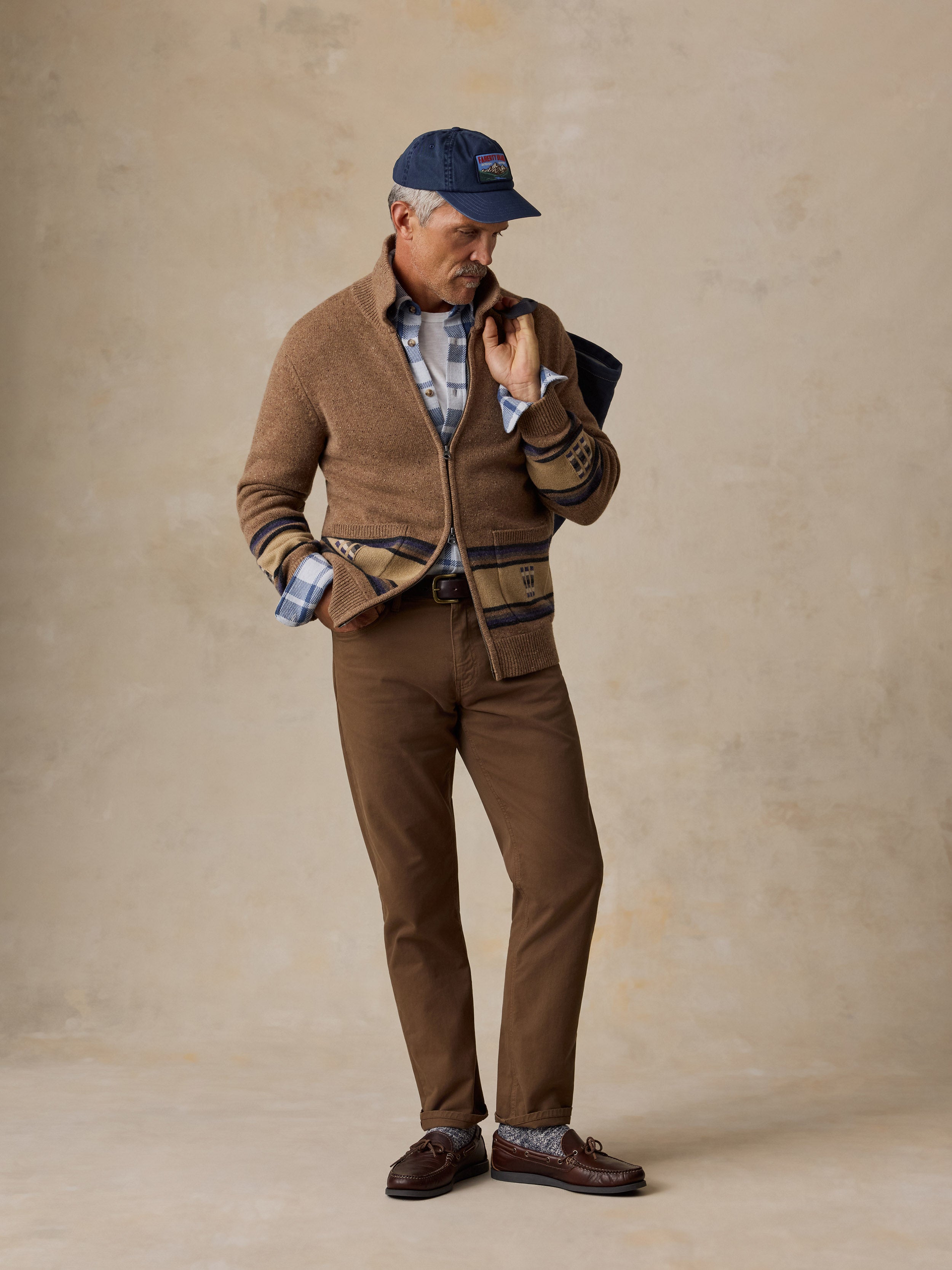 A man stands against a textured beige studio backdrop wearing a brown knit zip-front cardigan with striped trim, brown trousers, a blue-and-white plaid shirt, brown loafers, and a navy baseball cap.