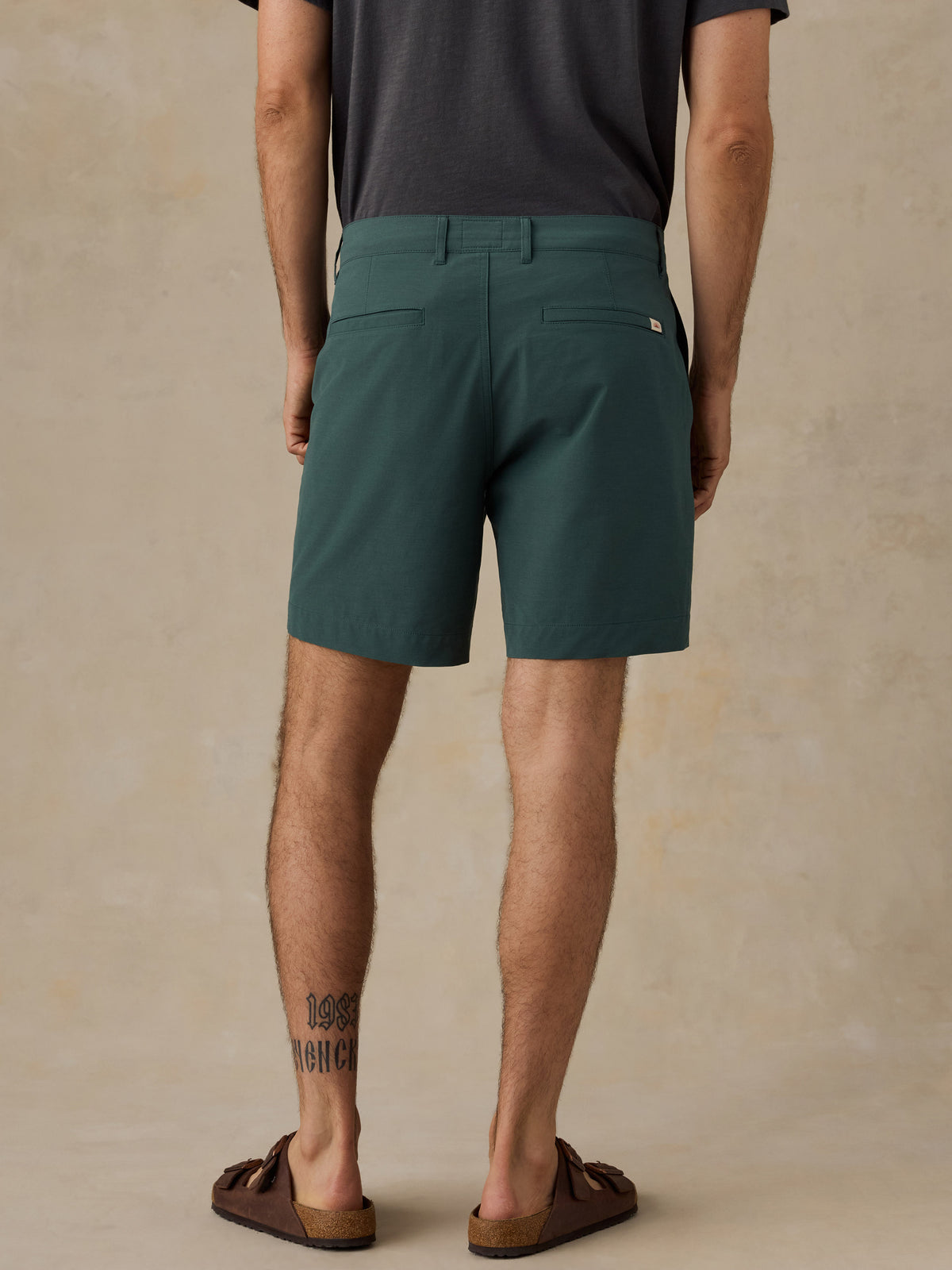 A man viewed from the back wearing dark teal-green shorts, a dark gray t-shirt, and brown slide sandals, standing against a textured beige studio backdrop., image 3 of 5