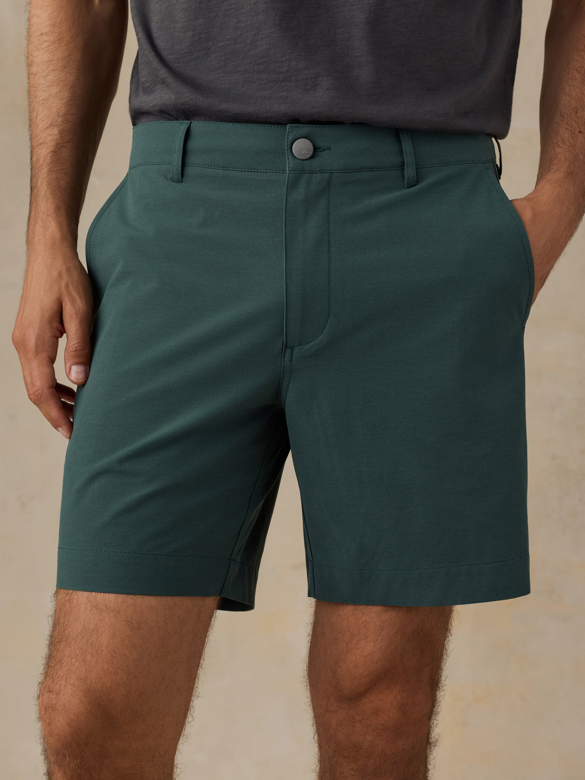A close-up of a person wearing dark teal-green tailored shorts with a button closure and belt loops, paired with a dark gray shirt against a warm beige textured background., image 4 of 5