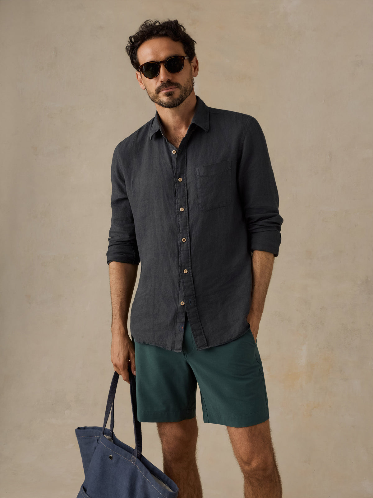 A man wearing a dark charcoal gray linen button-down shirt, teal green shorts, and sunglasses, holding a navy blue tote bag against a textured beige background., image 5 of 5