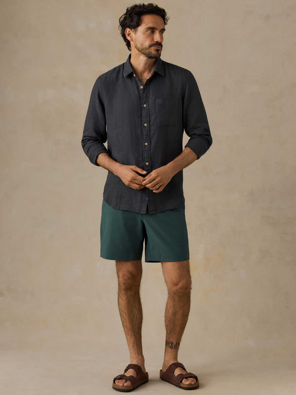A man wearing a dark charcoal gray long-sleeve button-down shirt, teal green shorts, and brown leather slide sandals, standing against a textured beige studio backdrop., image 2 of 5
