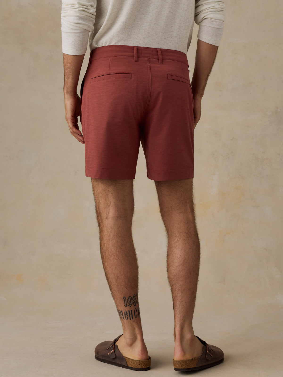 A man viewed from the back wearing rust-red chino shorts, a cream long-sleeve shirt, and brown leather slide sandals against a textured beige studio backdrop., image 3 of 6