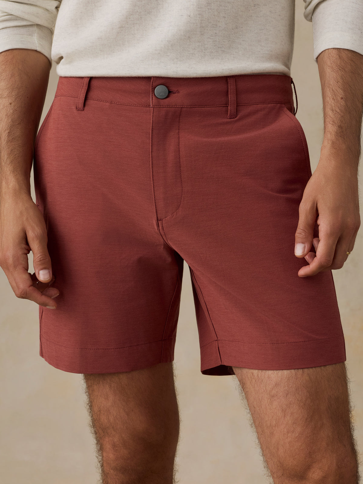 A close-up of a man wearing rust-colored chino shorts with a cream knit top, shown from the waist to mid-thigh against a warm beige background., image 4 of 6