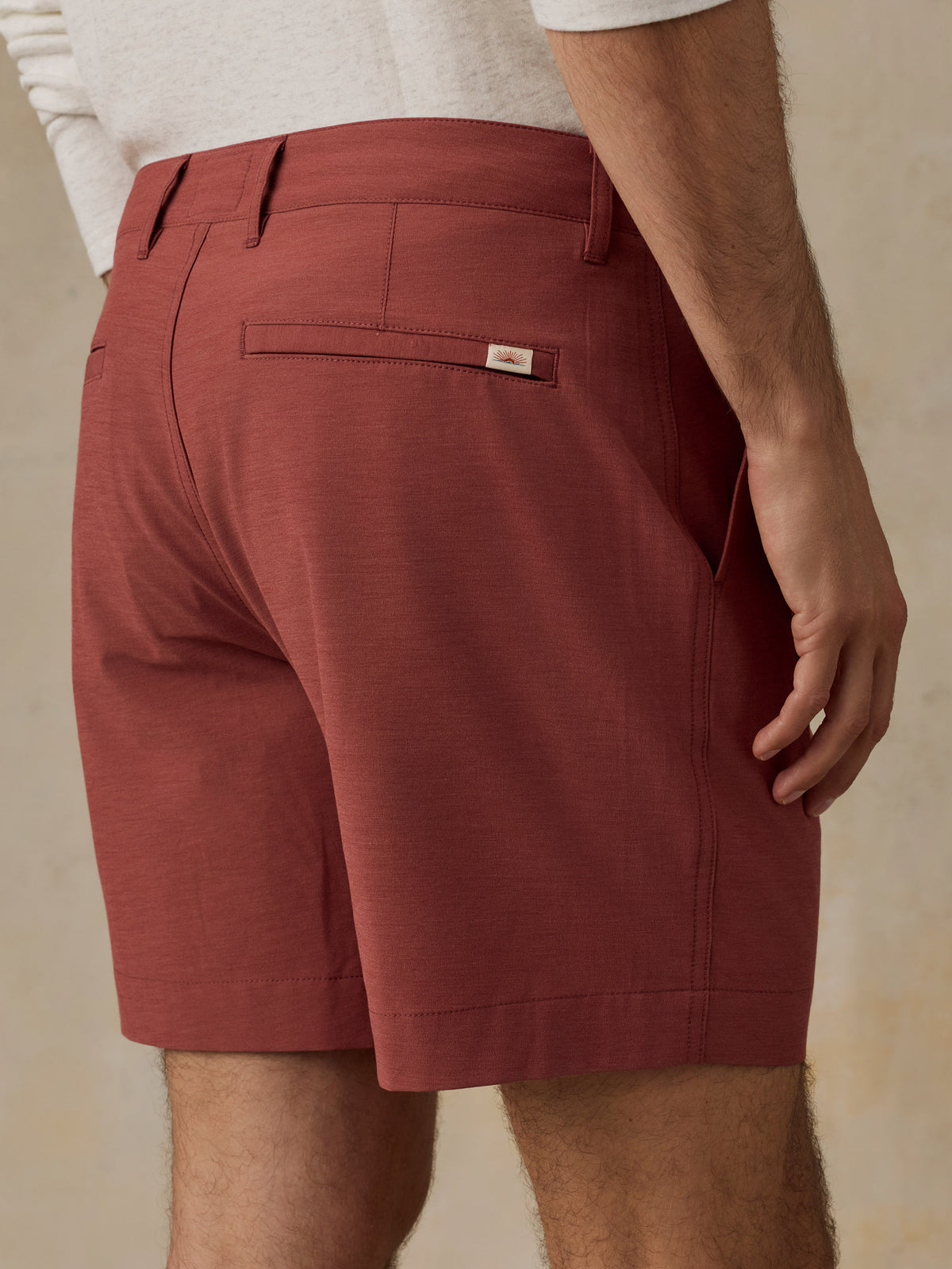 A close-up back view of a man wearing rust-colored chino shorts with a white shirt partially visible at the top, set against a neutral beige background., image 6 of 6