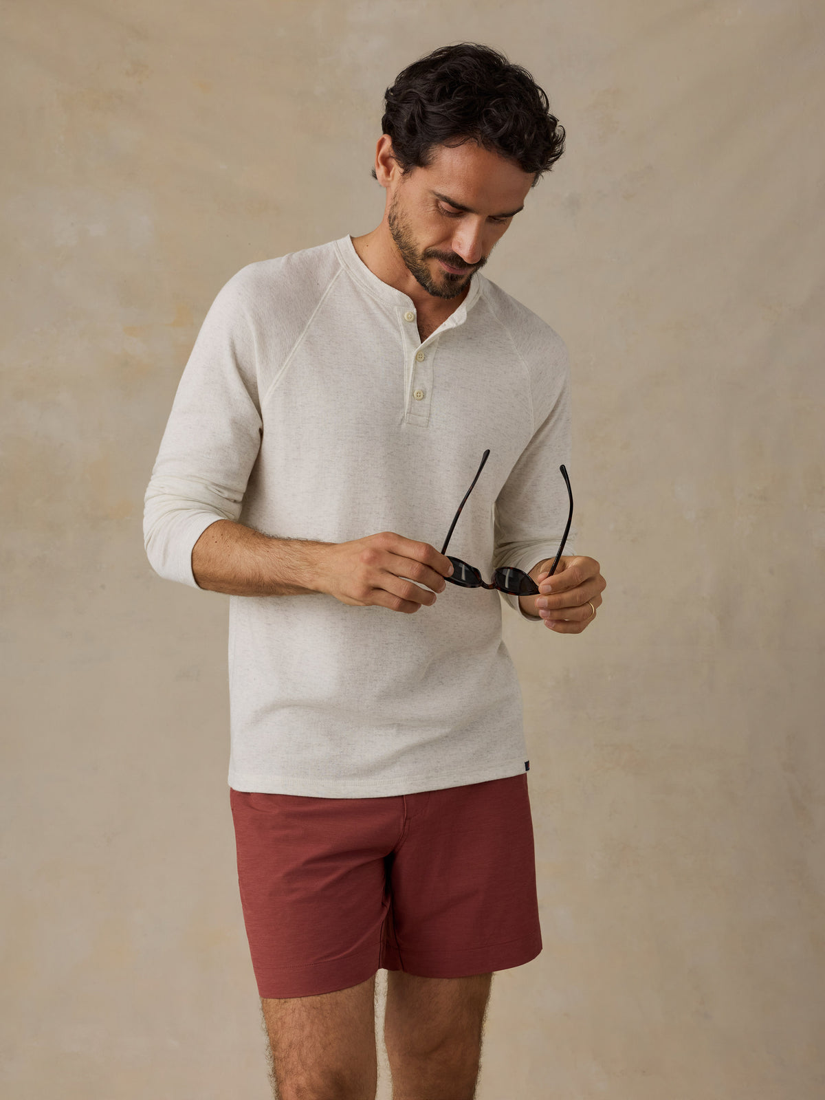 A man wearing a light beige long-sleeve henley shirt and rust-colored shorts, holding a pair of black sunglasses against a textured beige background., image 5 of 6
