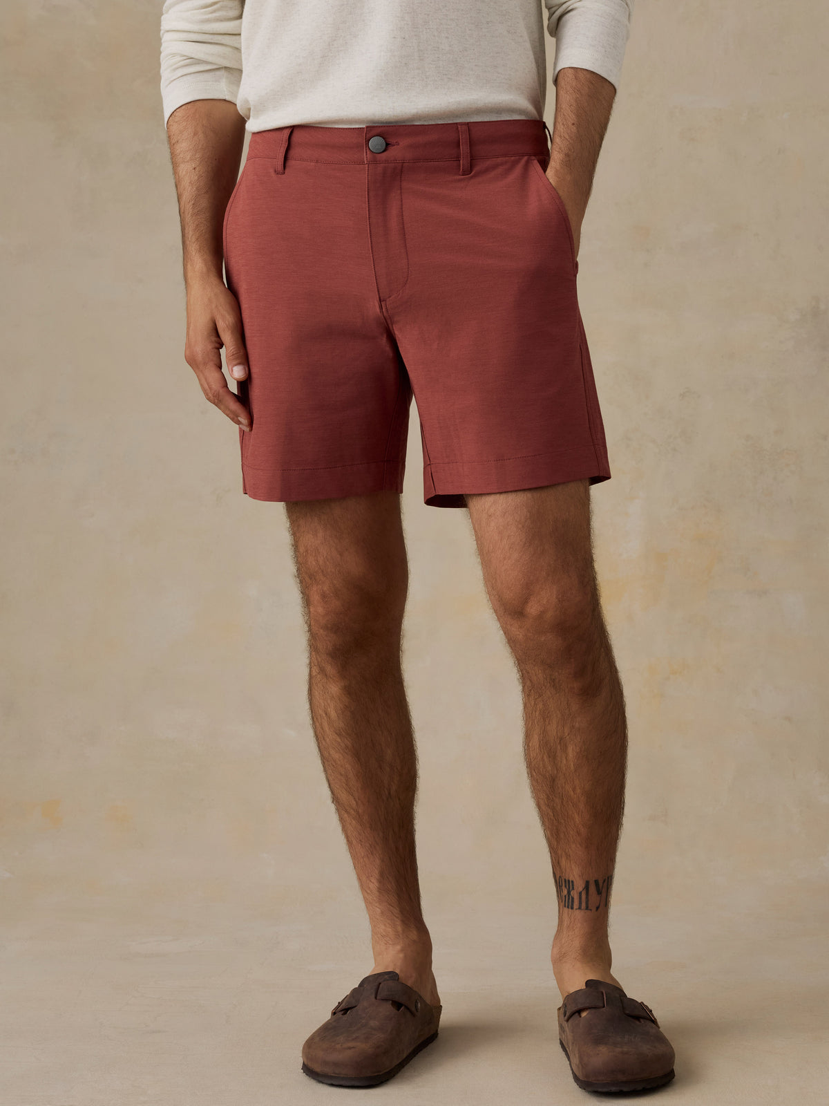 A man wearing rust-colored chino shorts, a cream long-sleeve shirt, and brown suede mules, standing against a textured beige studio backdrop., image 1 of 6
