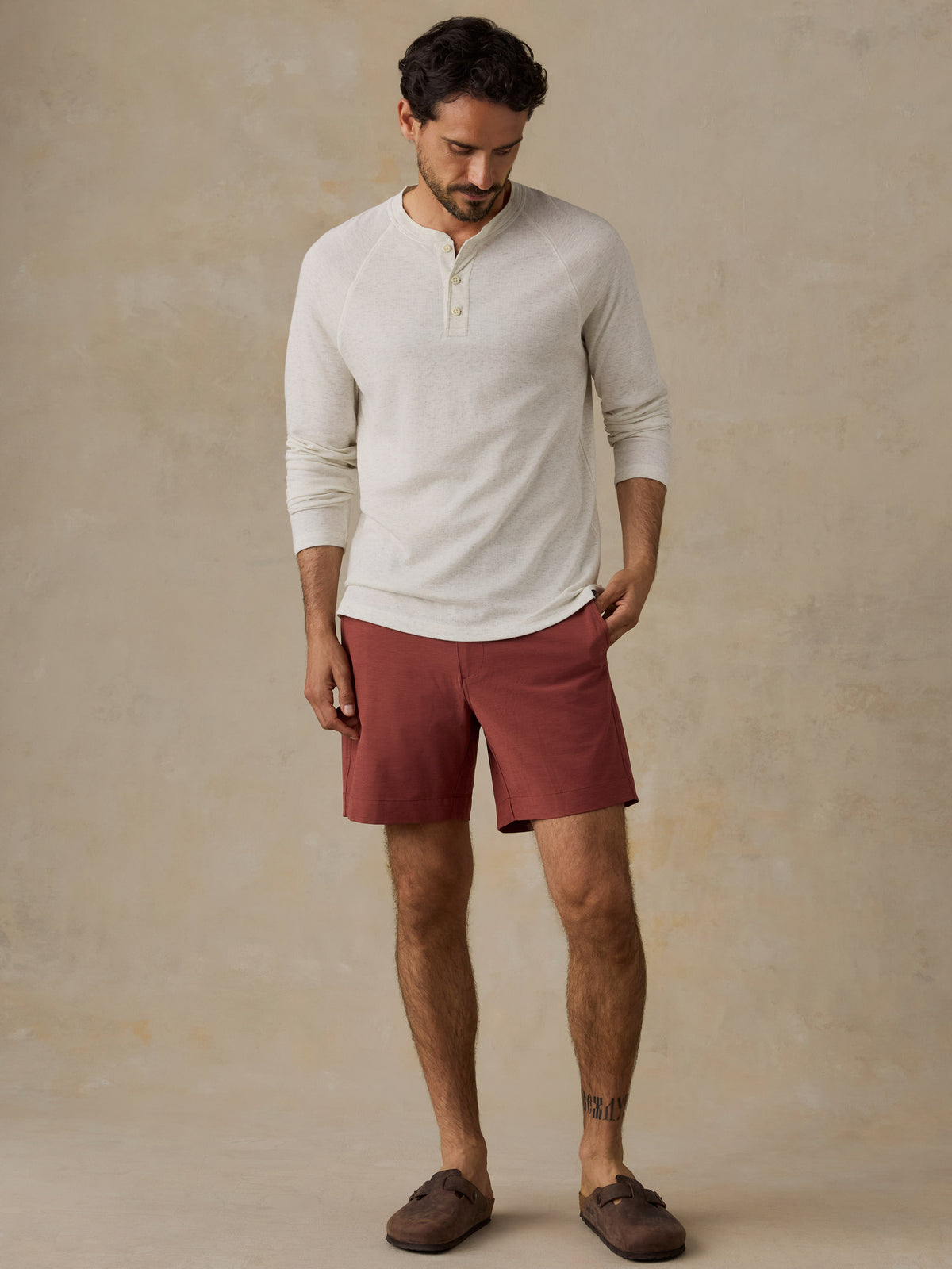 A man wearing a cream-colored long-sleeve henley shirt and rust-red shorts, standing against a textured beige studio backdrop., image 2 of 6