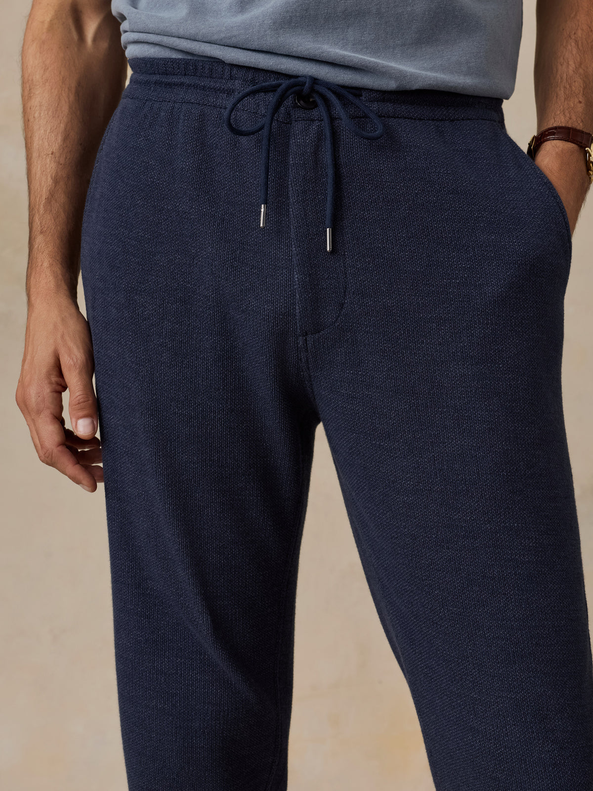 A close-up of a man wearing dark navy knit lounge pants with a drawstring waistband, paired with a light blue-gray top. The photo is cropped from the chest down against a warm beige studio background., image 4 of 6