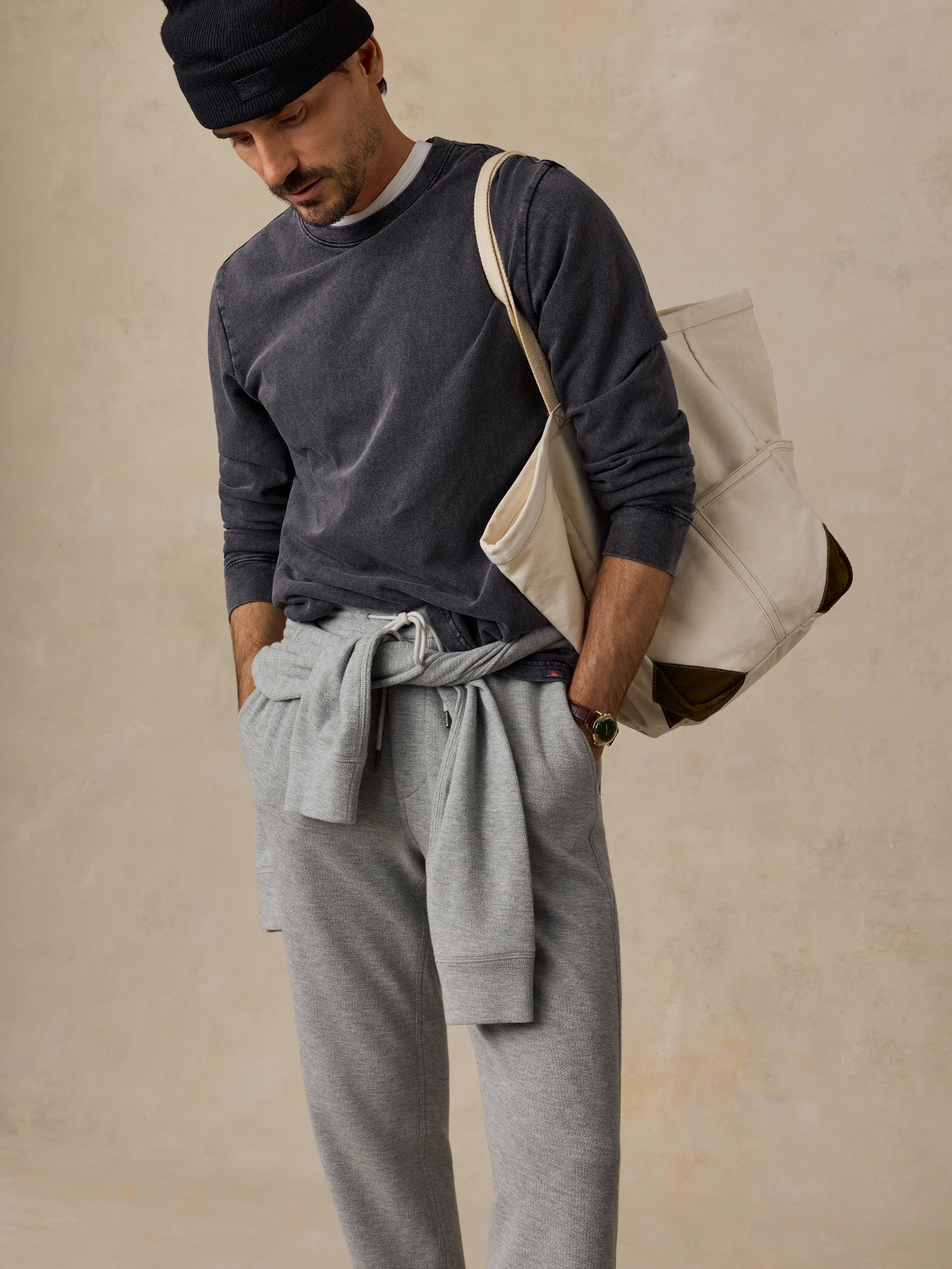 A man wearing a dark gray long-sleeve shirt, light gray sweatpants, a black beanie, and carrying a large cream canvas tote bag over his shoulder, set against a textured beige wall.