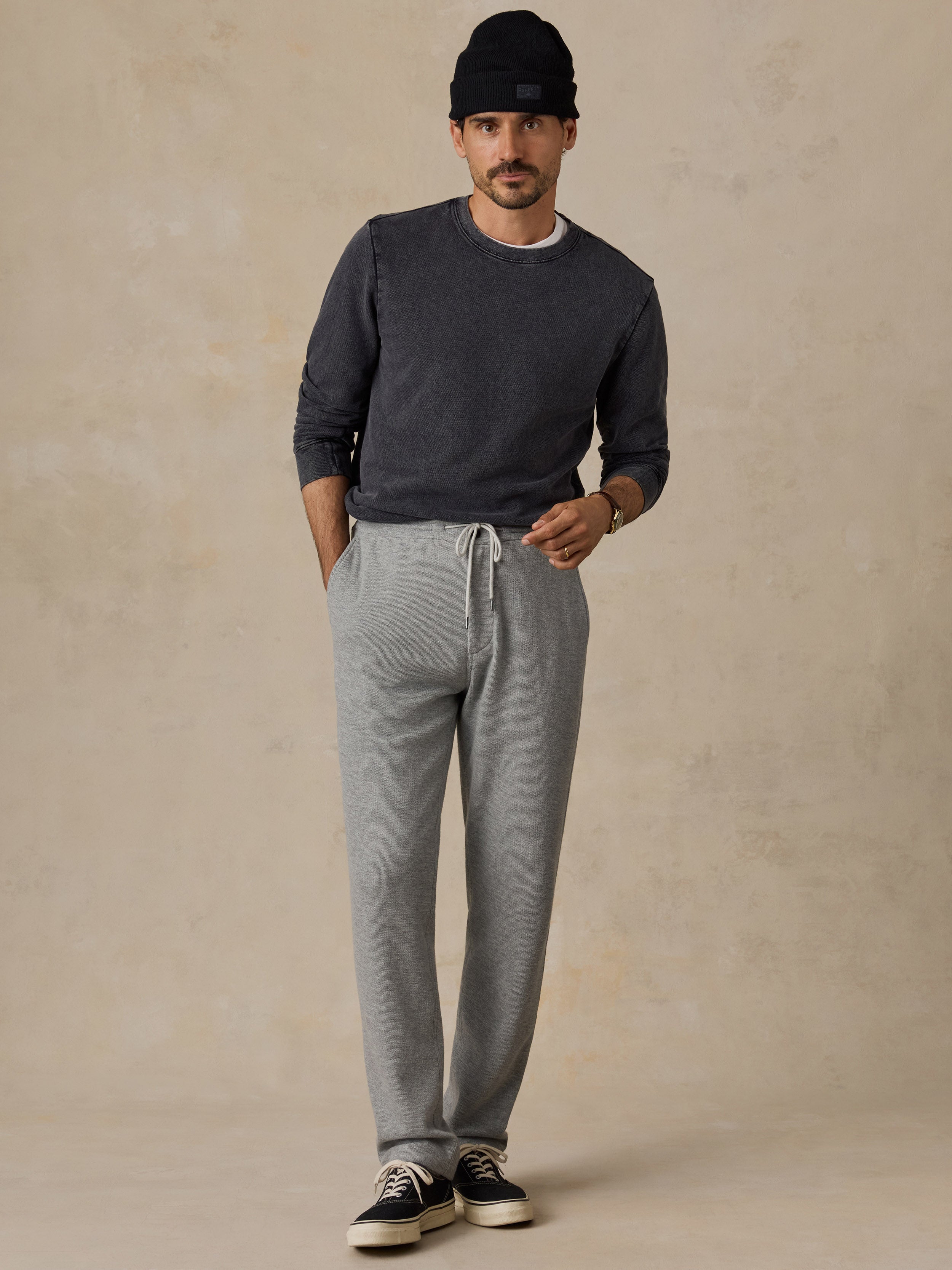 A man wearing a dark gray long-sleeve crewneck sweater, light gray heather jogger sweatpants, a black beanie, and black sneakers, standing against a textured beige studio backdrop.