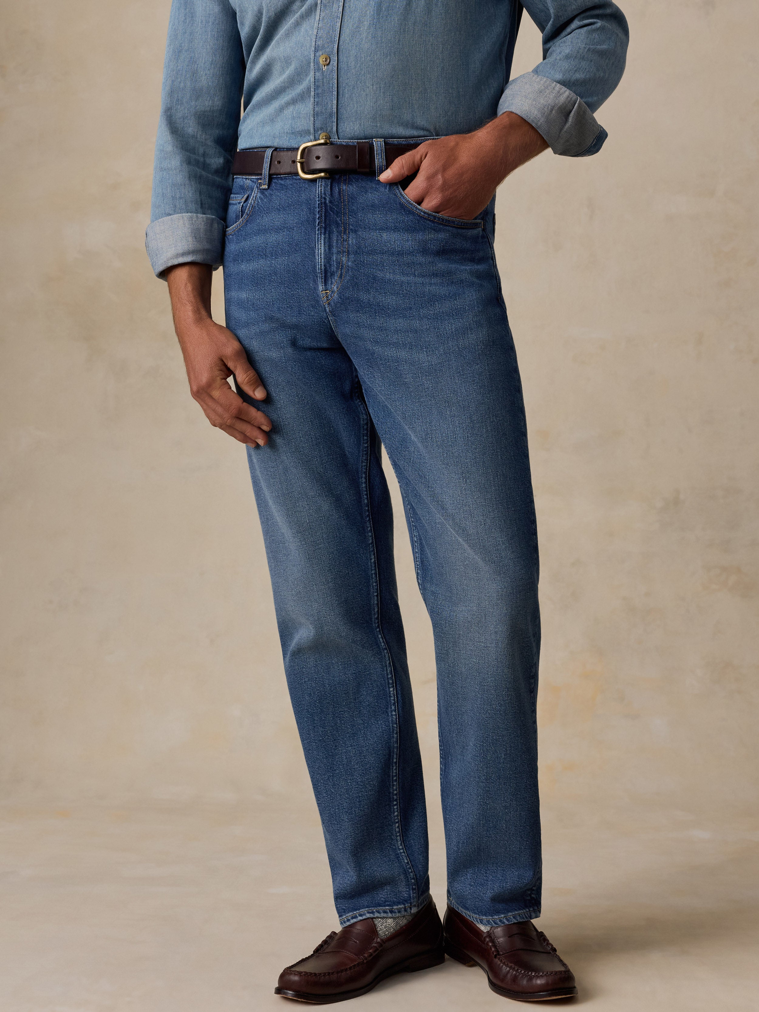 A person wearing medium-wash blue jeans with a brown belt, a light blue denim shirt, and brown leather loafers, standing against a textured beige studio backdrop.