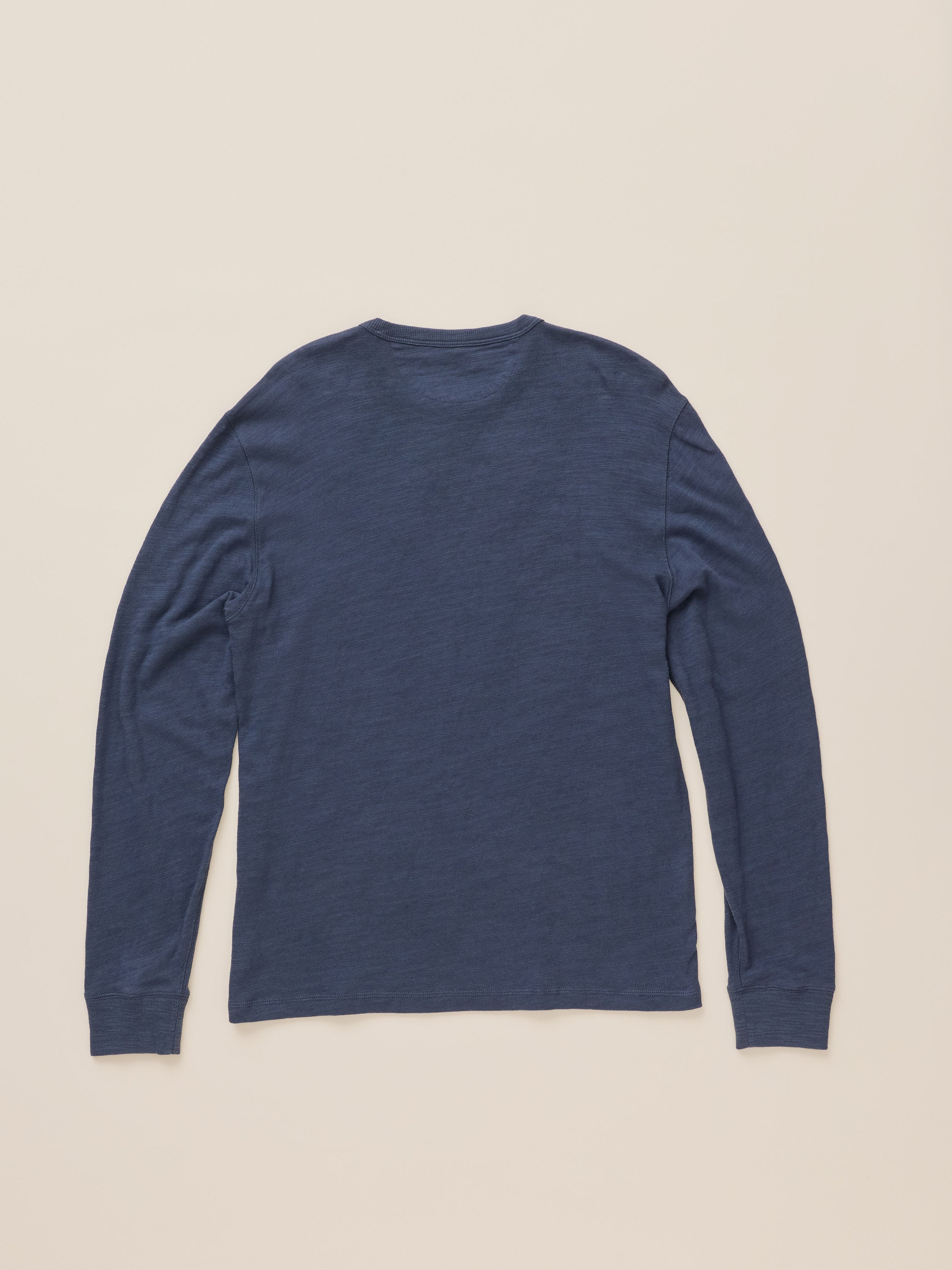 A navy blue long-sleeve crewneck shirt shown from the back, laid flat against a solid light beige background.