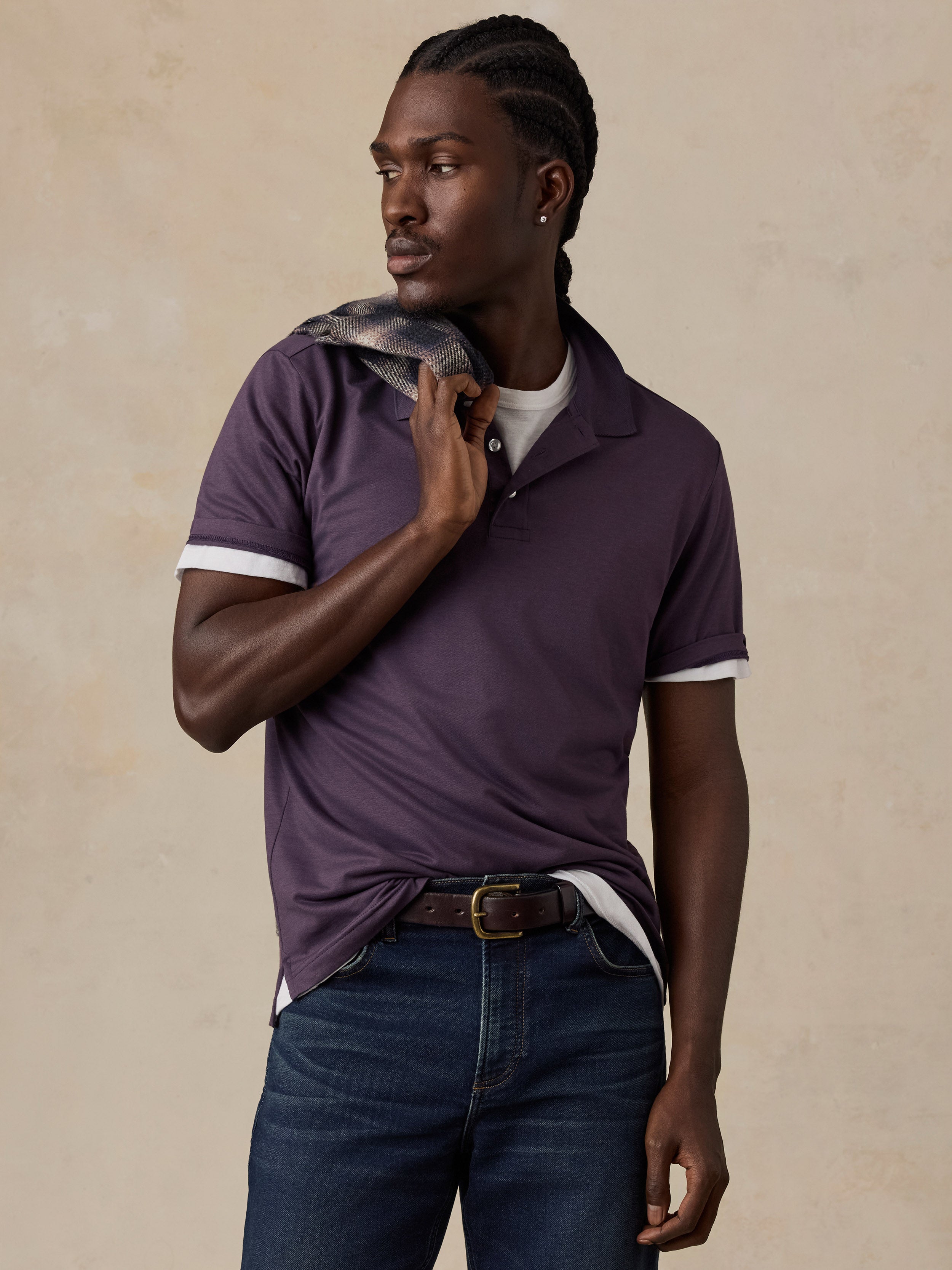 A man wearing a dark purple polo shirt, styled with dark blue jeans and a brown belt, standing against a textured beige background.