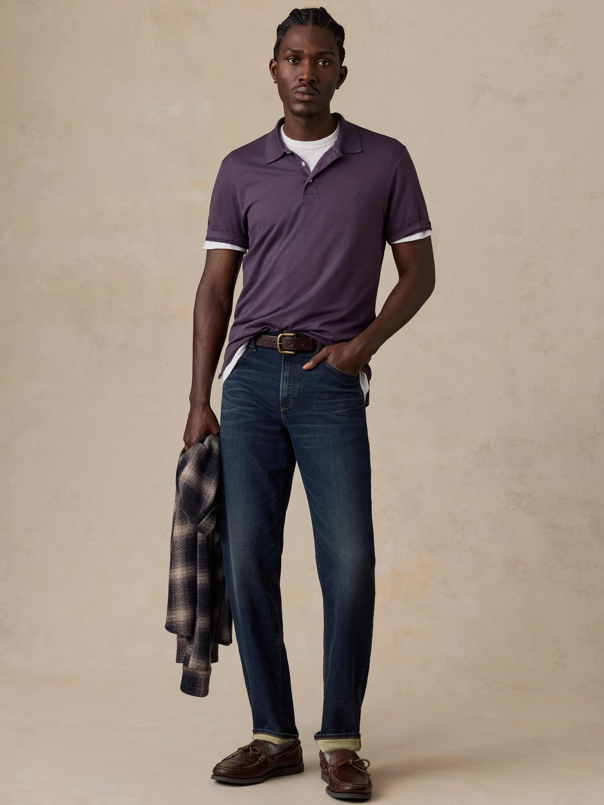 A man modeling a short-sleeve polo shirt in a deep plum purple color with white trim on the collar and sleeve cuffs, paired with dark blue jeans, brown leather loafers, and holding a plaid shirt under his arm, set against a textured beige studio backdrop.