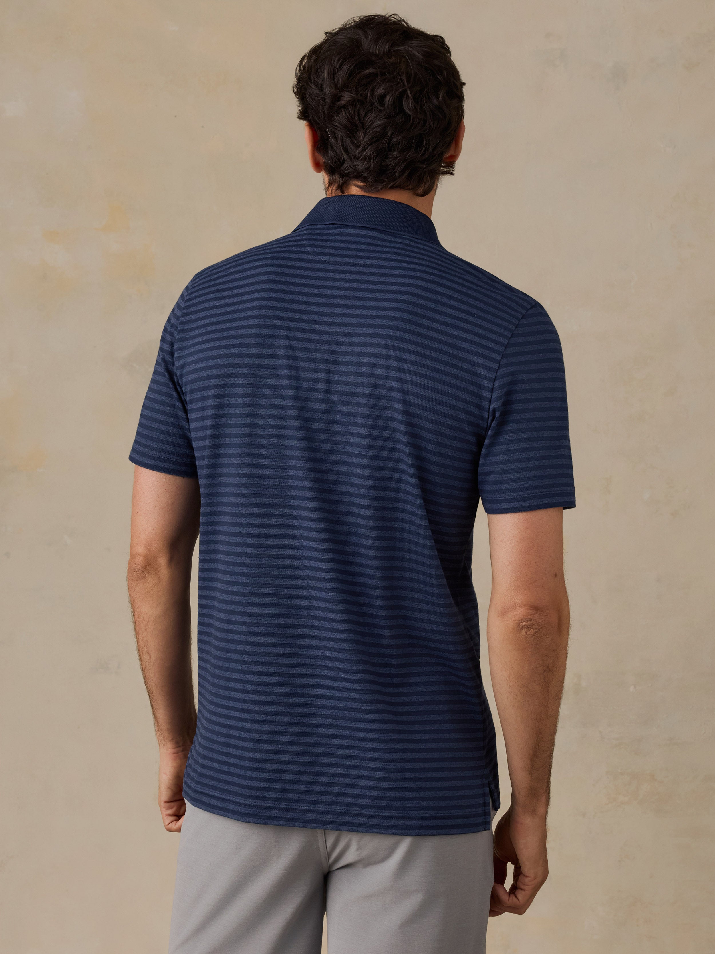 A man viewed from the back wearing a navy blue short-sleeve polo shirt with thin horizontal stripes and light gray pants, standing against a textured beige wall.
