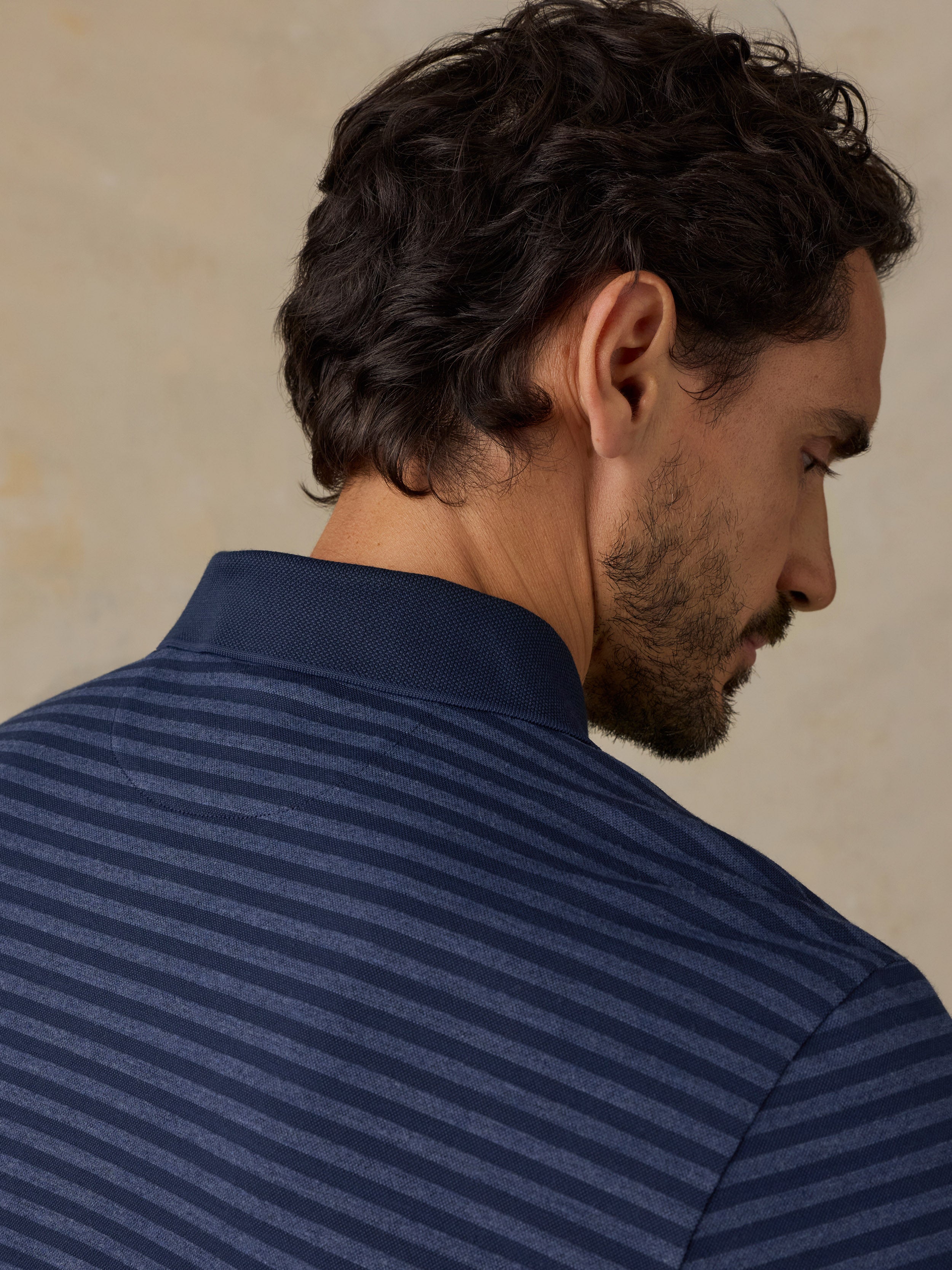 A close-up side profile of a man wearing a navy blue polo shirt with fine horizontal stripes, set against a neutral beige background.
