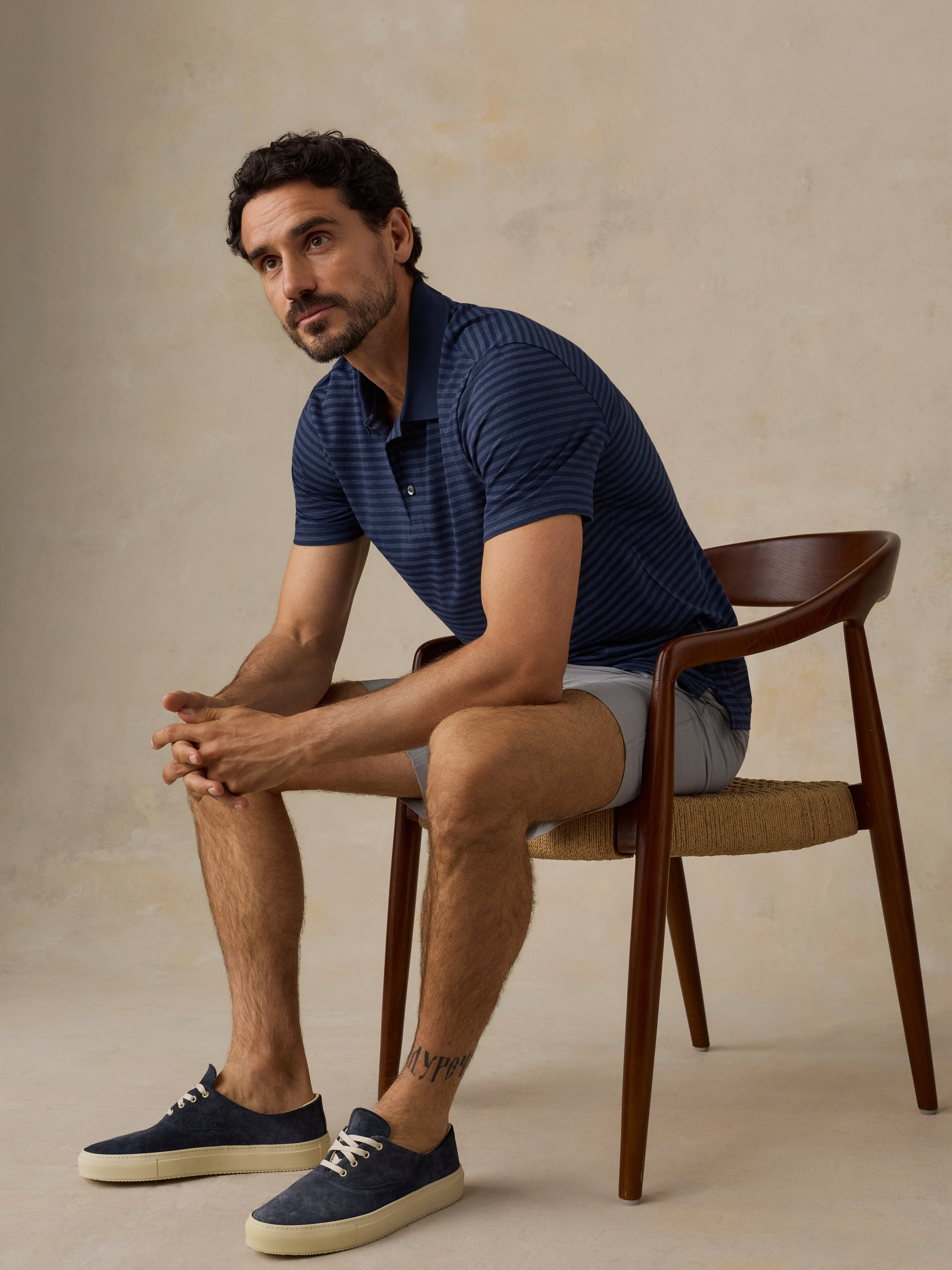 A man sits on a wooden chair wearing a navy blue striped short-sleeve polo shirt, light gray shorts, and navy blue slip-on shoes with cream soles, set against a textured beige studio background.