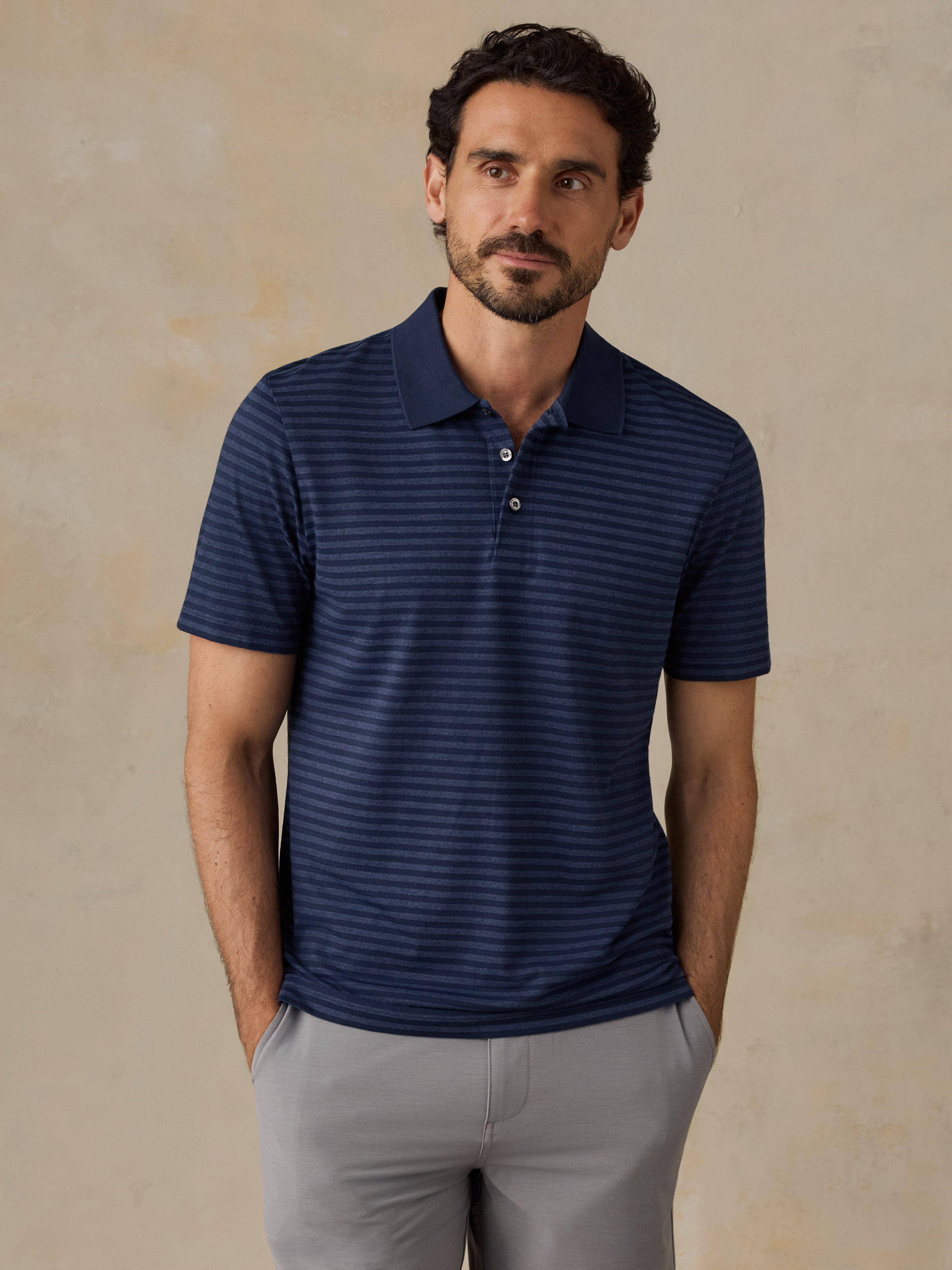 A man wearing a navy blue short-sleeve polo shirt with thin horizontal stripes, paired with light gray trousers, standing against a textured beige background.