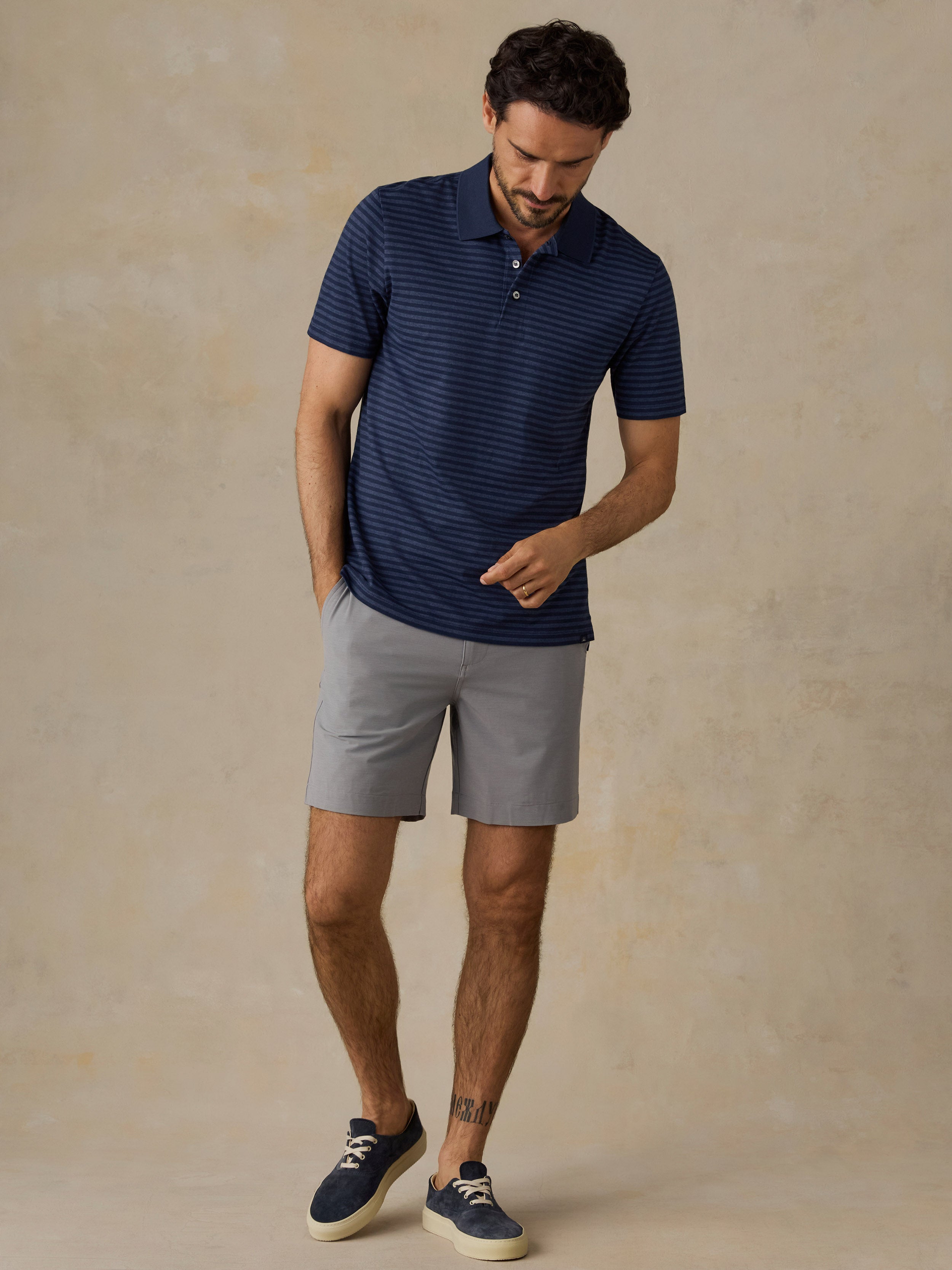 A man wearing a navy blue short-sleeve polo shirt with thin horizontal stripes, light gray shorts, and navy blue sneakers, standing against a textured beige studio backdrop.