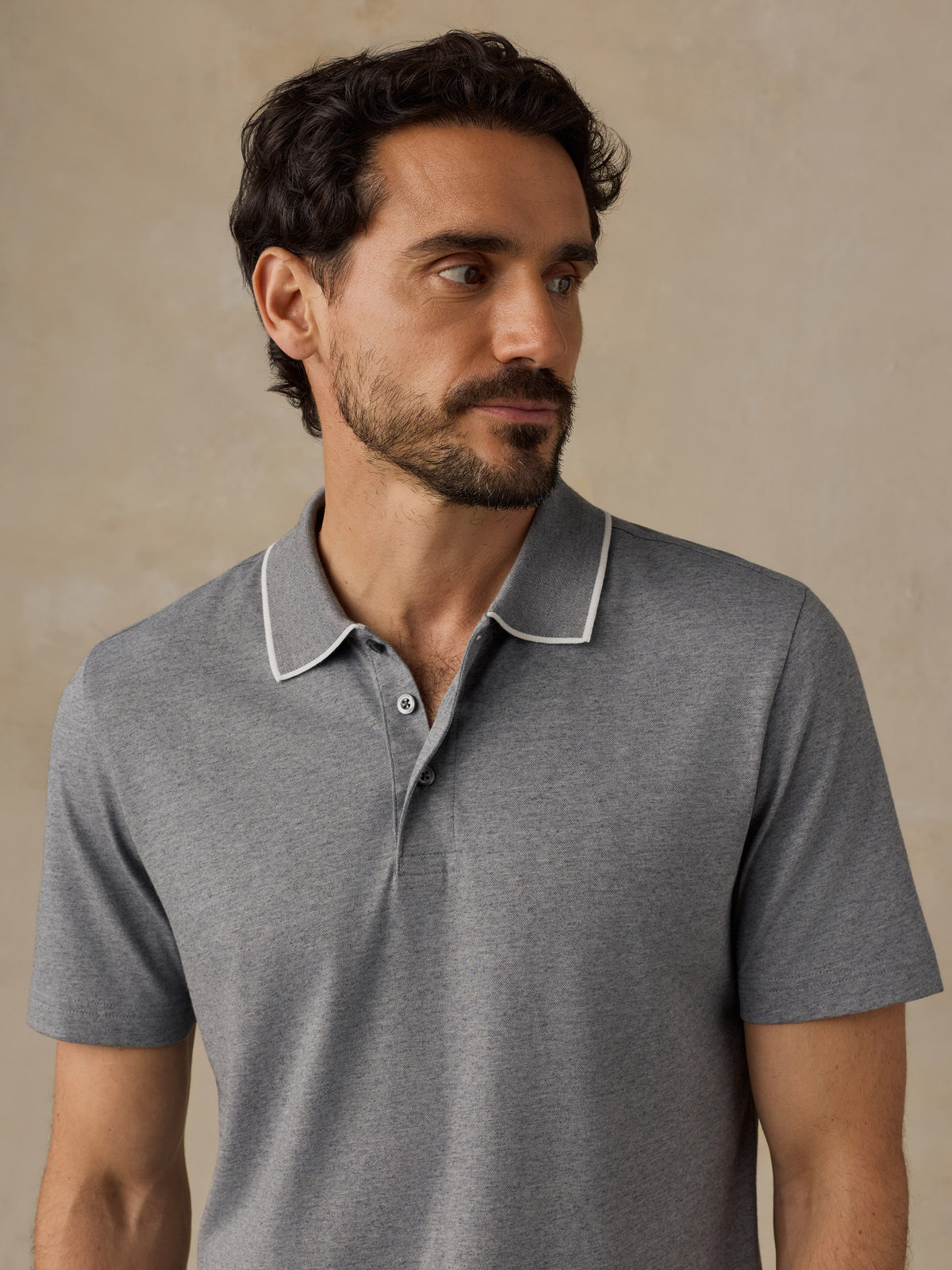 A man wearing a heather gray short-sleeve polo shirt with a white contrast piped collar, shown from the chest up against a warm beige textured background., image 5 of 6