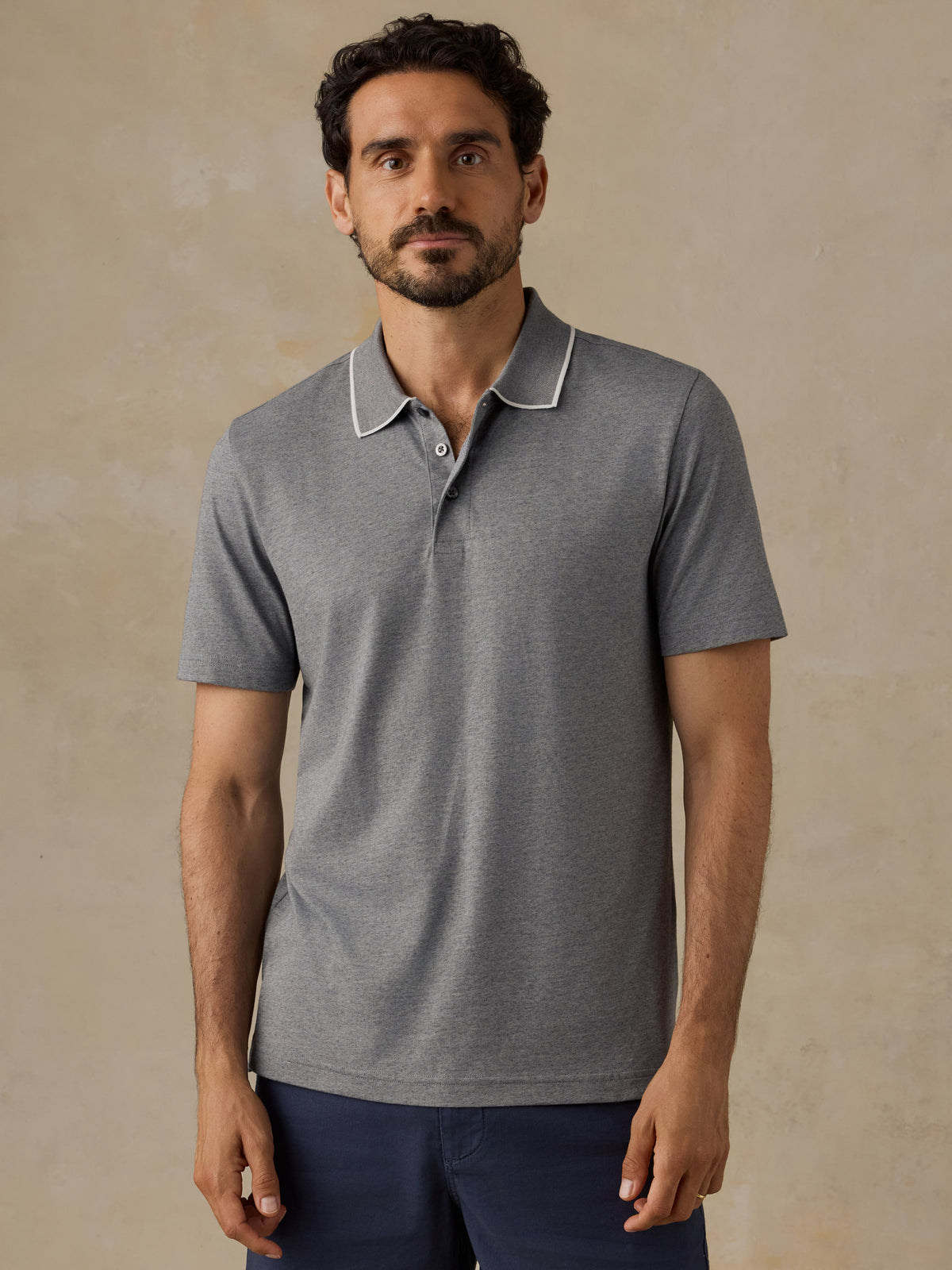 A man wearing a heather gray short-sleeve polo shirt with a white contrast collar, paired with navy blue pants, standing against a textured beige background., image 1 of 6