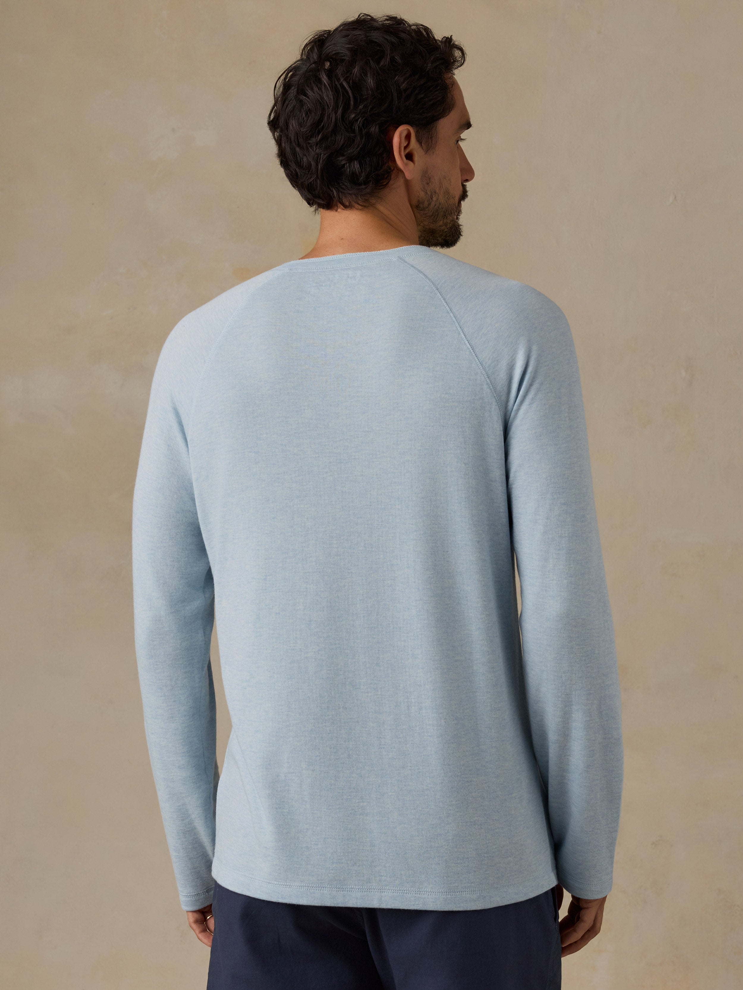 A man viewed from the back wearing a light blue long-sleeve knit top and dark navy shorts, standing against a textured beige studio backdrop.