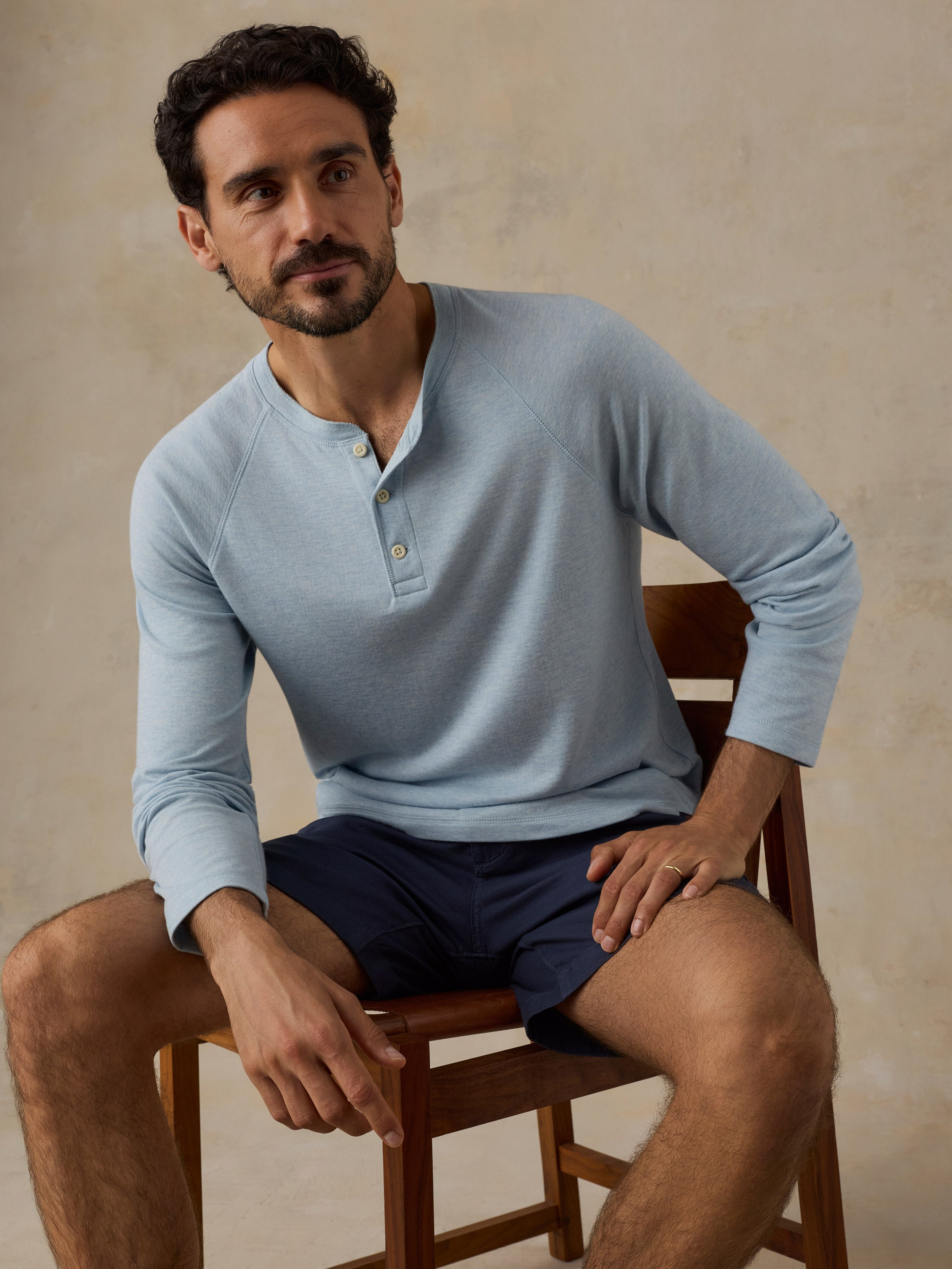 A man wearing a light blue long-sleeve henley shirt and navy shorts, sitting on a wooden chair against a textured beige wall.
