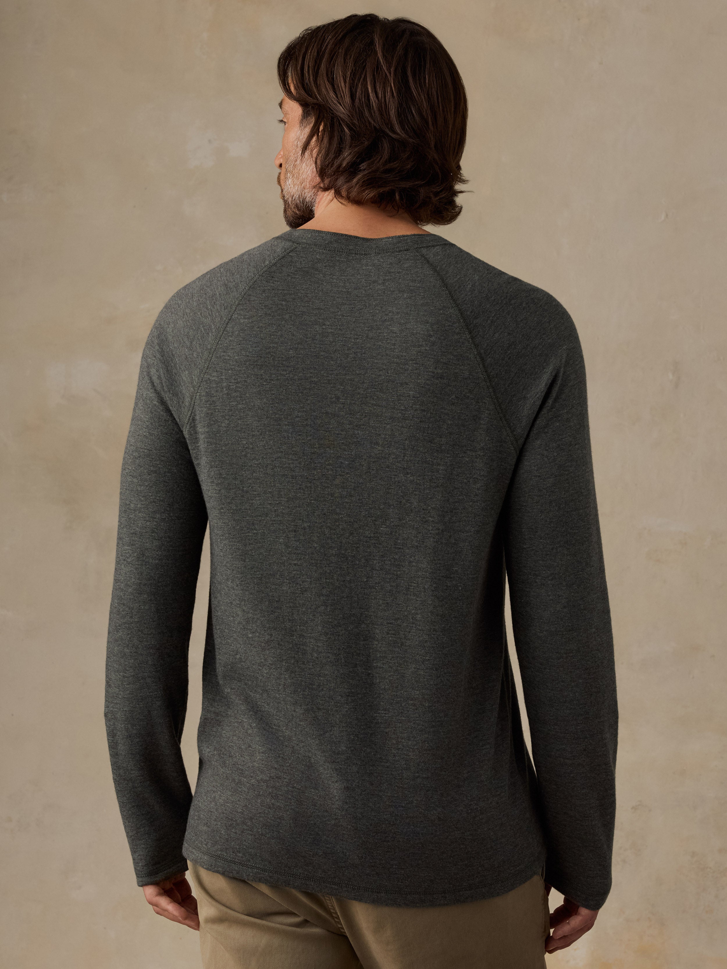 A man viewed from the back wearing a dark heather green long-sleeve raglan shirt, paired with tan pants, against a textured beige studio backdrop.