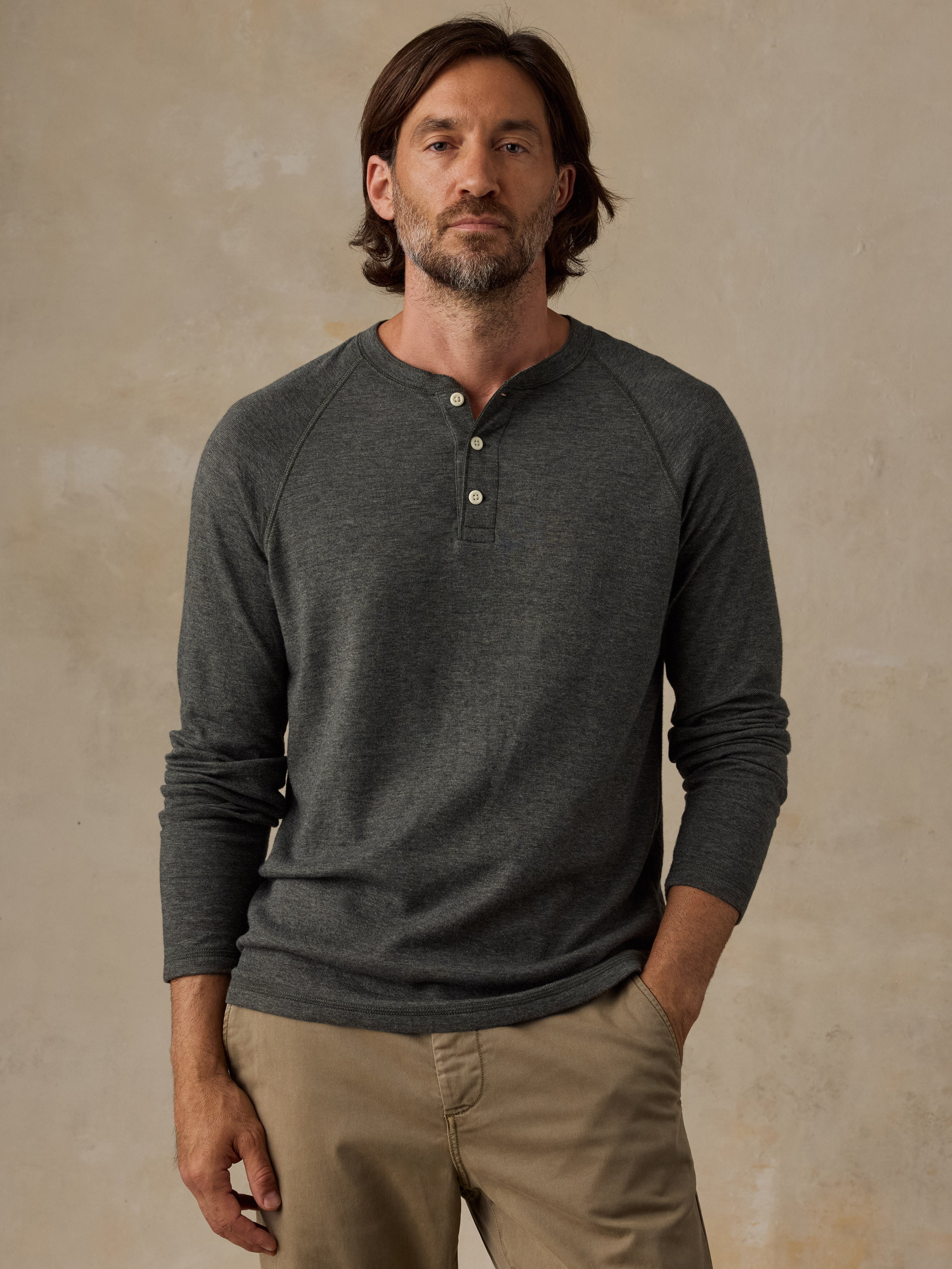 A man wearing a dark green long-sleeve henley shirt and khaki pants, standing against a textured beige studio background.