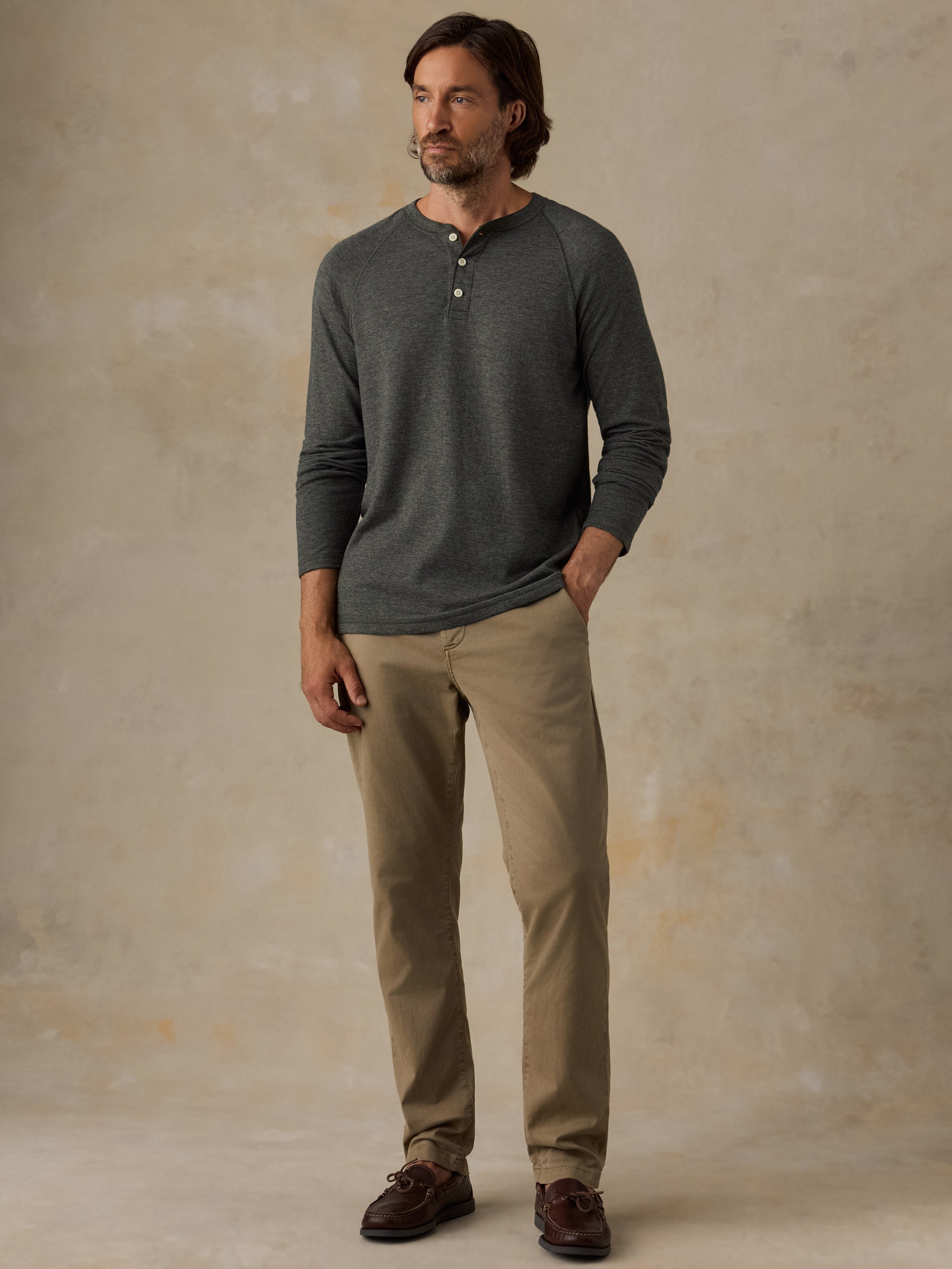 A man wearing a dark green long-sleeve henley shirt, tan chinos, and brown leather shoes, standing against a textured beige studio backdrop.