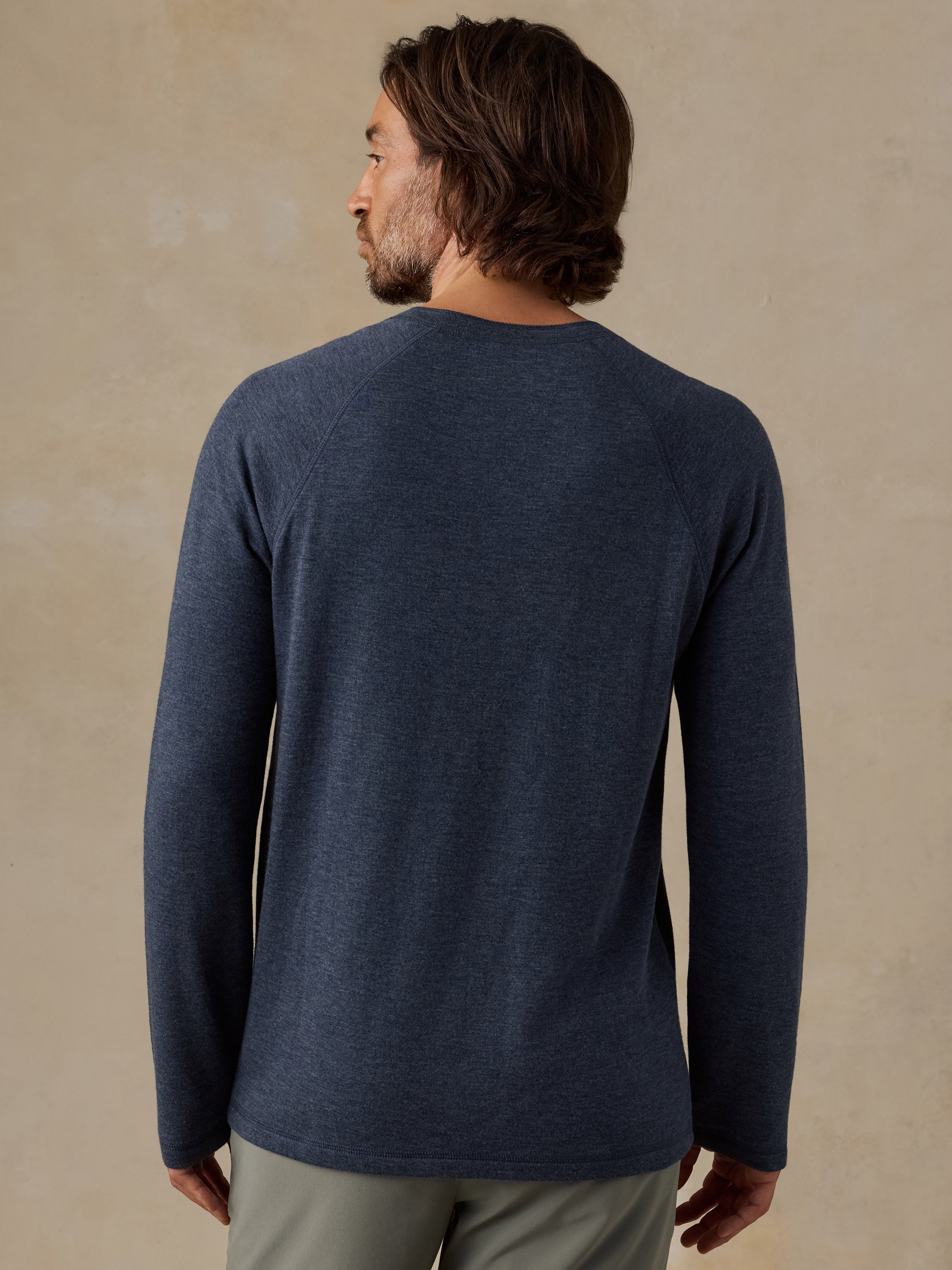 A man viewed from the back wearing a heathered navy blue long-sleeve shirt and olive green shorts, standing against a textured beige studio background.