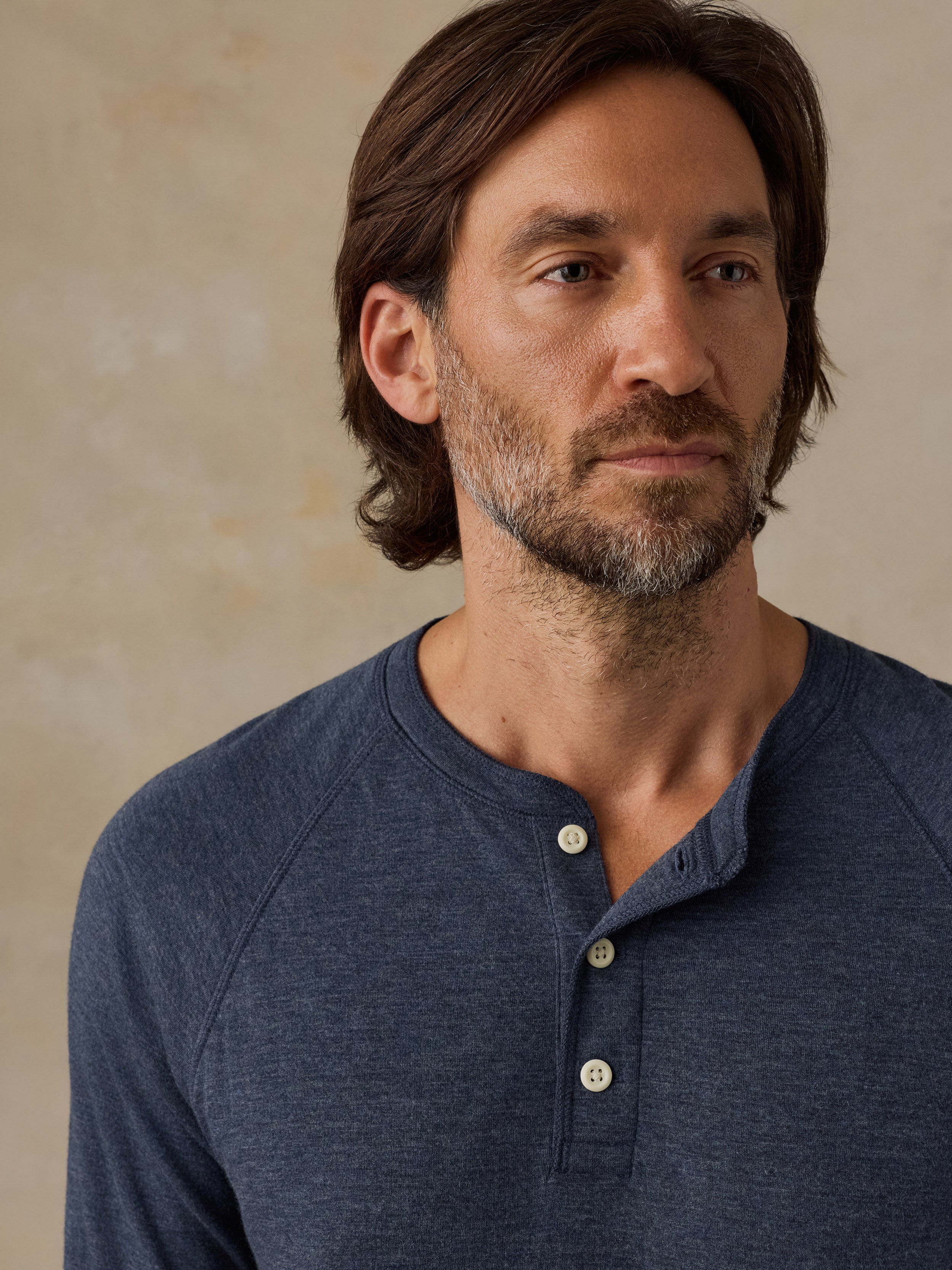 A man wearing a heathered navy blue henley shirt with three buttons, shown in a close-up portrait against a textured beige background.
