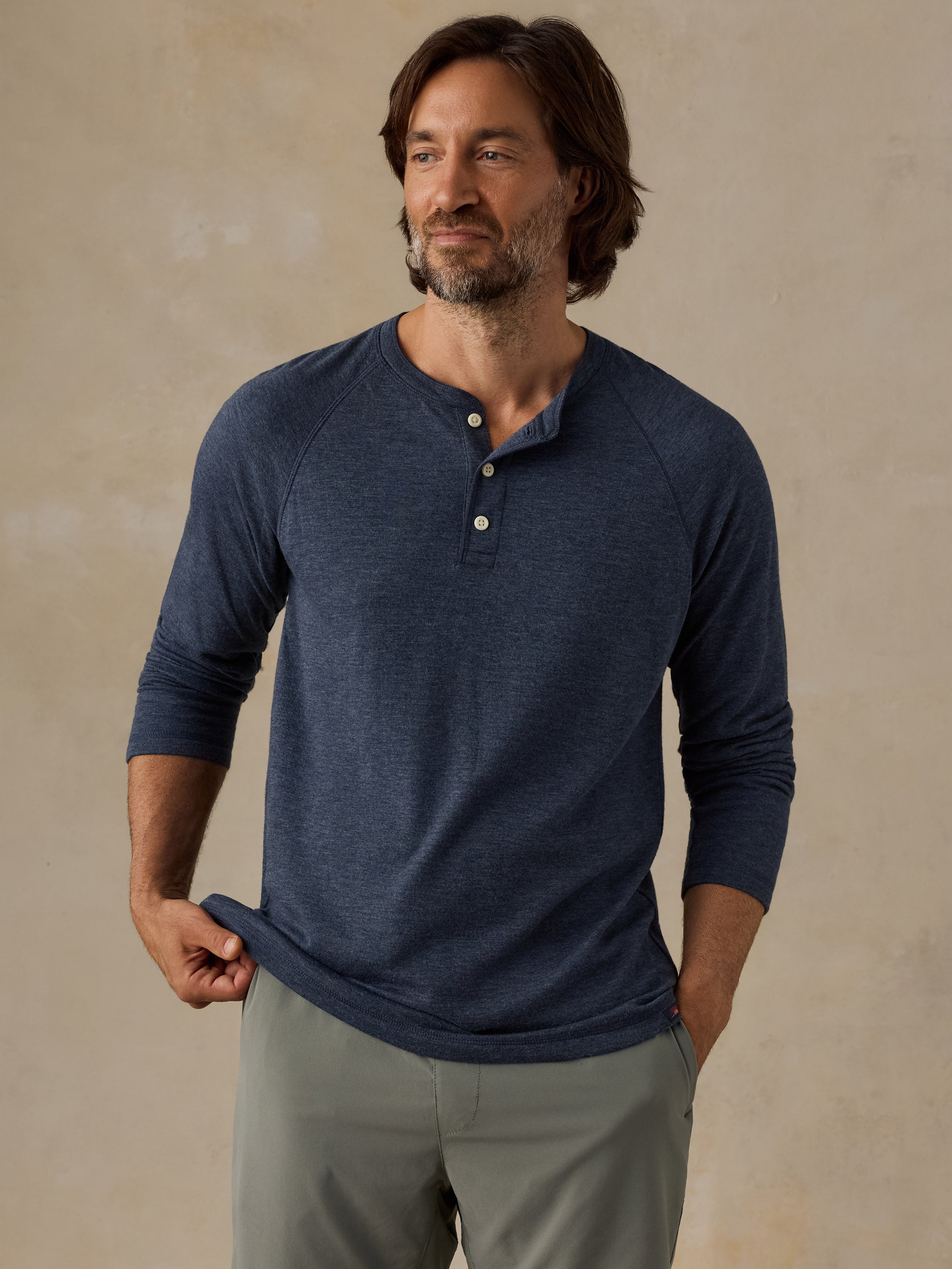 A man wearing a navy blue long-sleeve henley shirt with three buttons and olive green shorts, standing against a textured beige studio background.