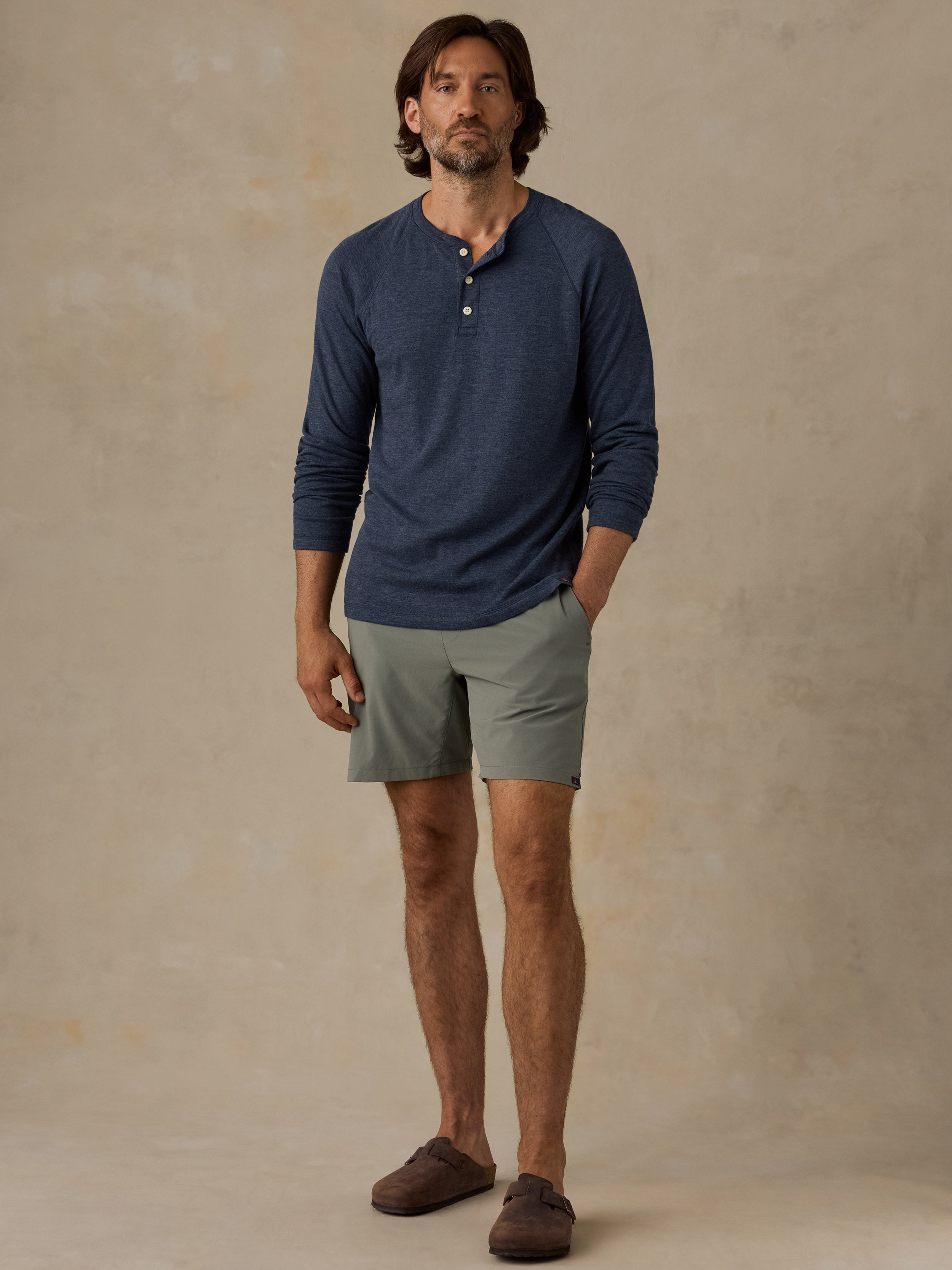 A man modeling a navy blue long-sleeve henley shirt and light olive green shorts against a textured beige studio backdrop.