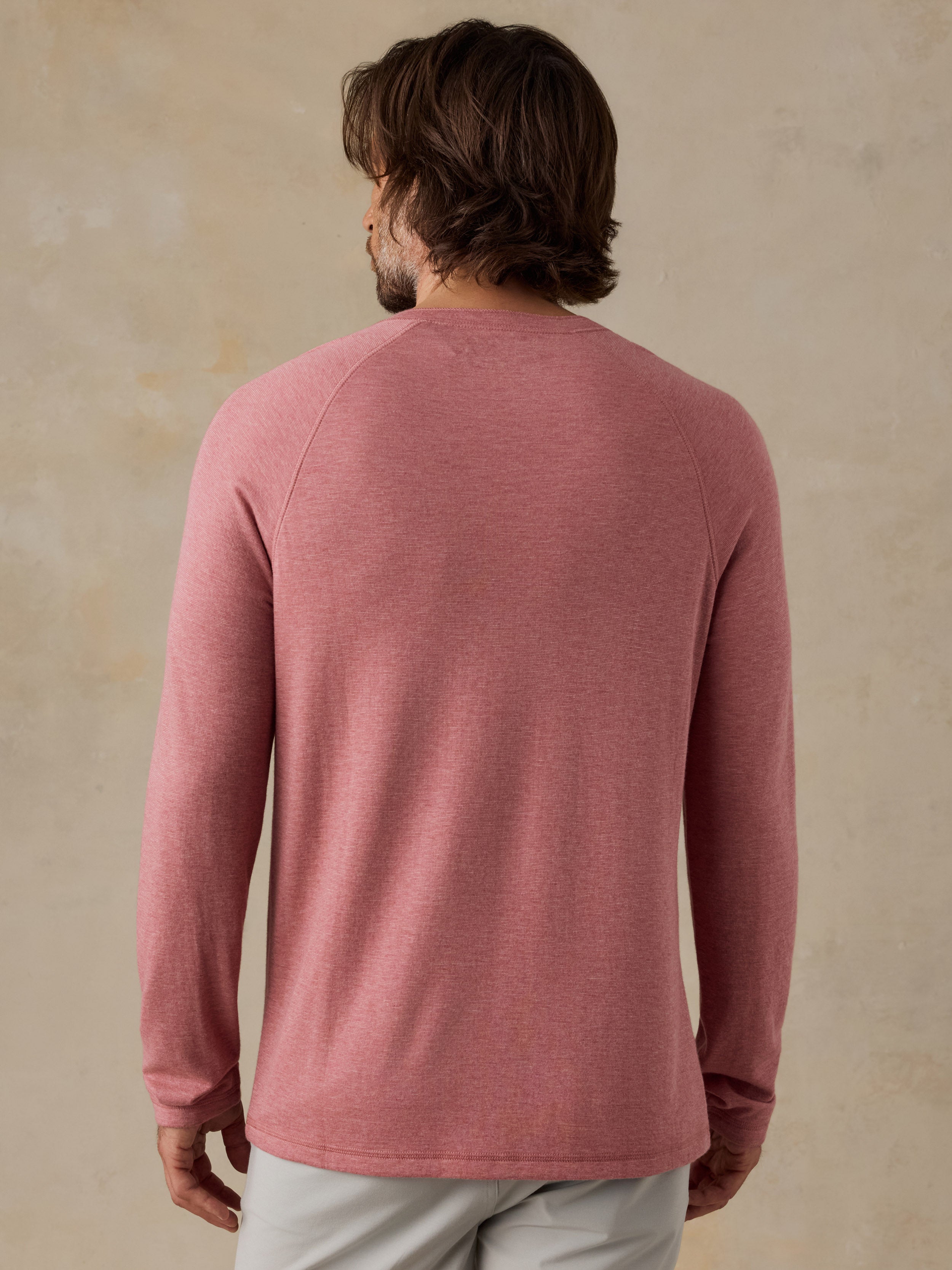 A man viewed from behind wearing a dusty rose long-sleeve henley shirt and light gray shorts, standing against a textured beige background.
