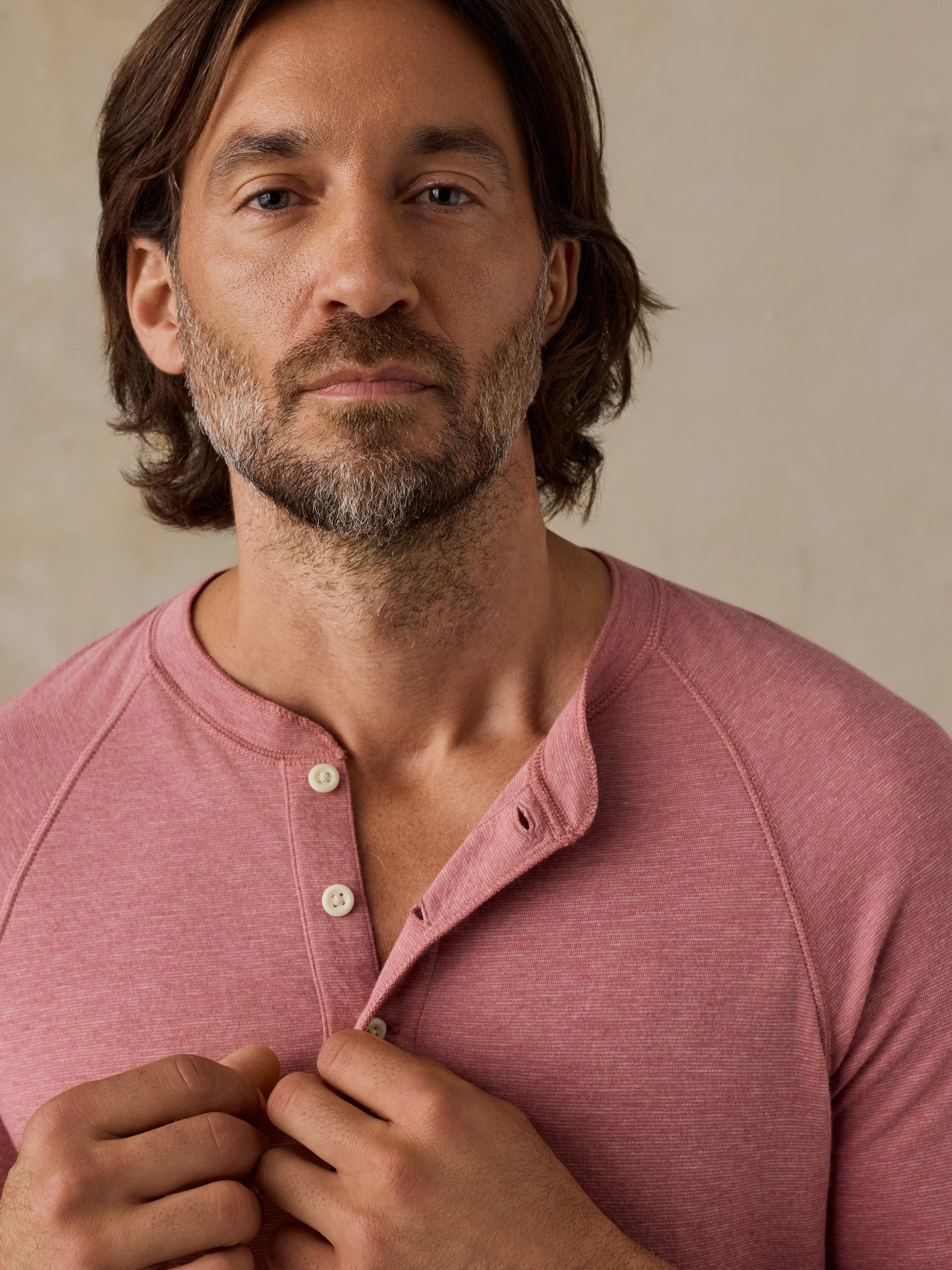 A close-up of a man wearing a dusty rose pink raglan henley shirt, buttoning the collar. The background is a soft, textured beige wall.